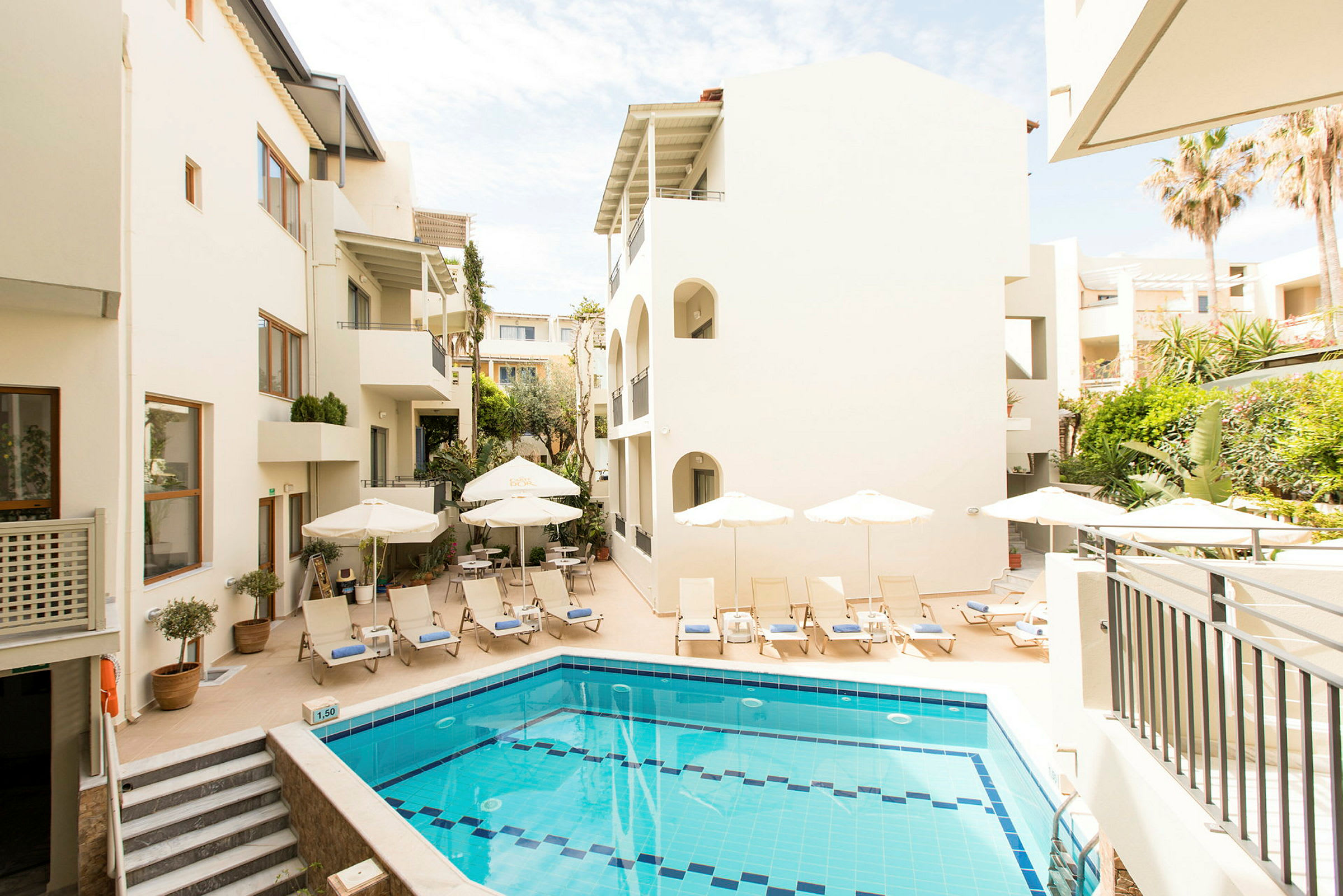 Hotellets poolområde med solstolar och parasoller, omgiven av byggnader och grönska. (AI-genererad text)