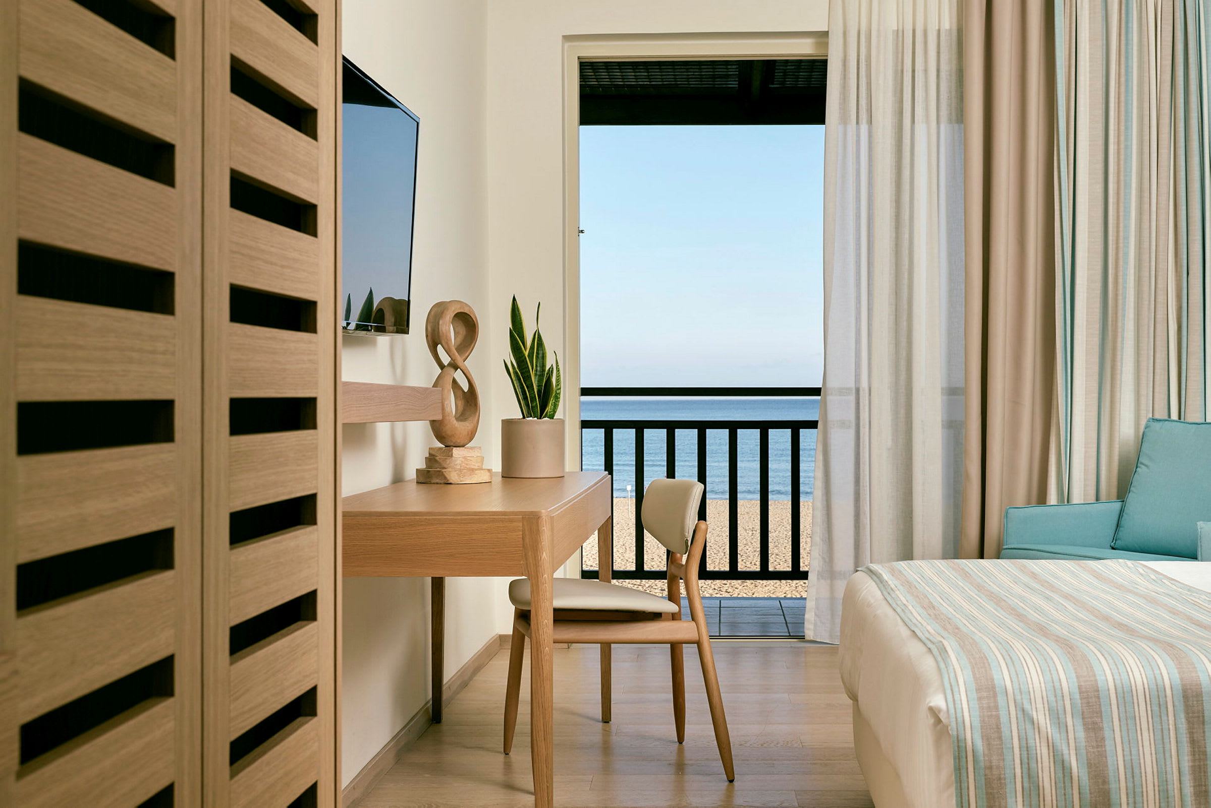 Hotellrum med skrivbord, stol och säng med utsikt mot havet från balkongen. (AI-genererad text)