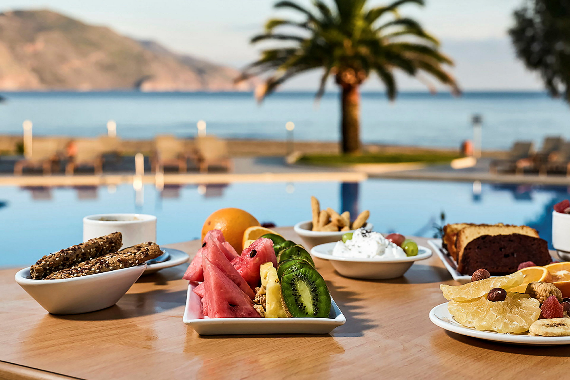 En frukost serveras vid ett bord nära en pool, med utsikt mot havet och palmträd. (AI-genererad text)