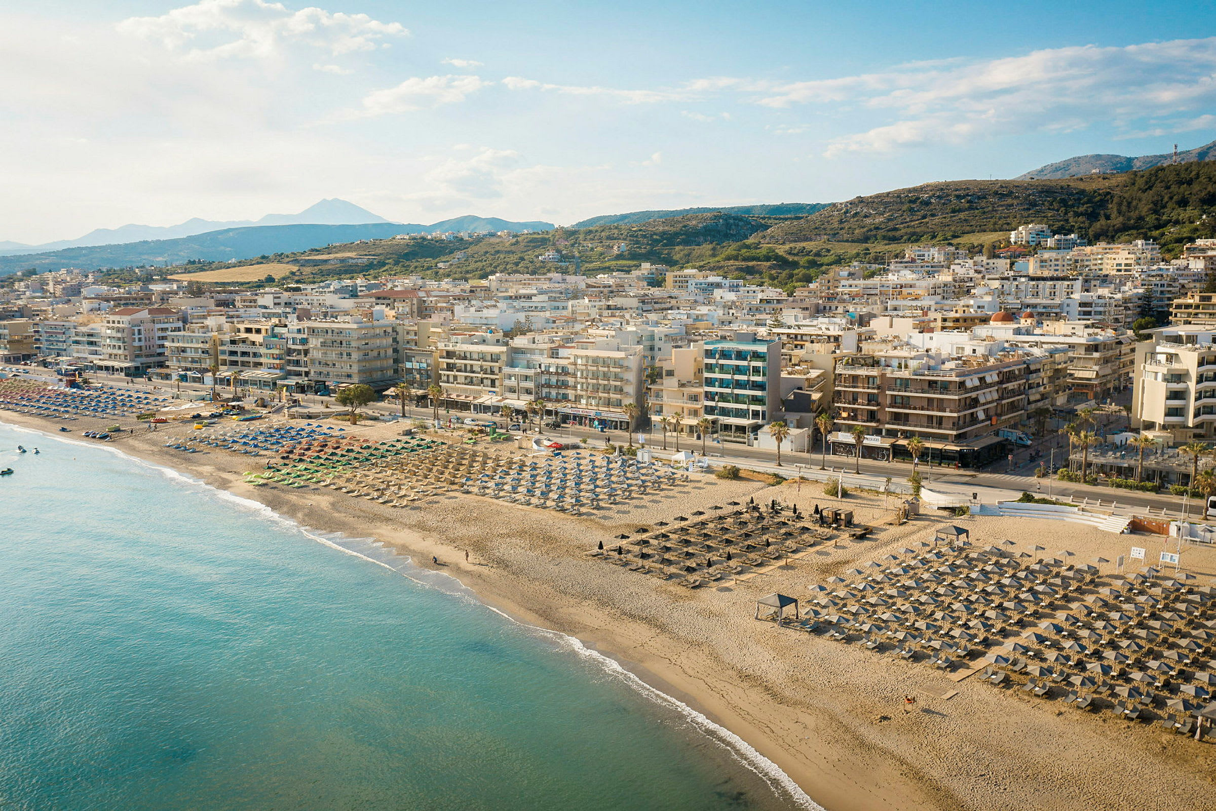 En sandstrand med parasoller och solstolar framför en stad med lägenhetsbyggnader och berg i bakgrunden. (AI-genererad text)