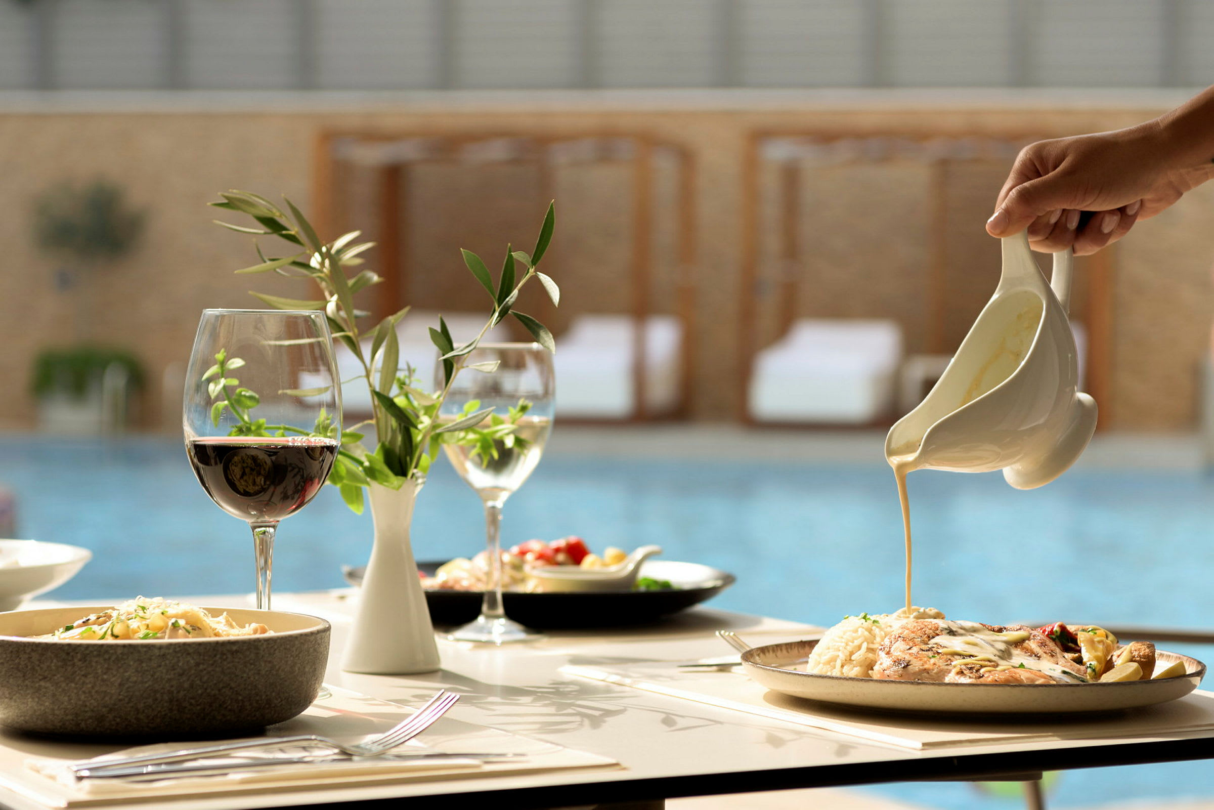En hand häller sås över en tallrik med maträtt på ett dukat bord med vin och dekorationer. Poolen syns i bakgrunden. (AI-genererad text)