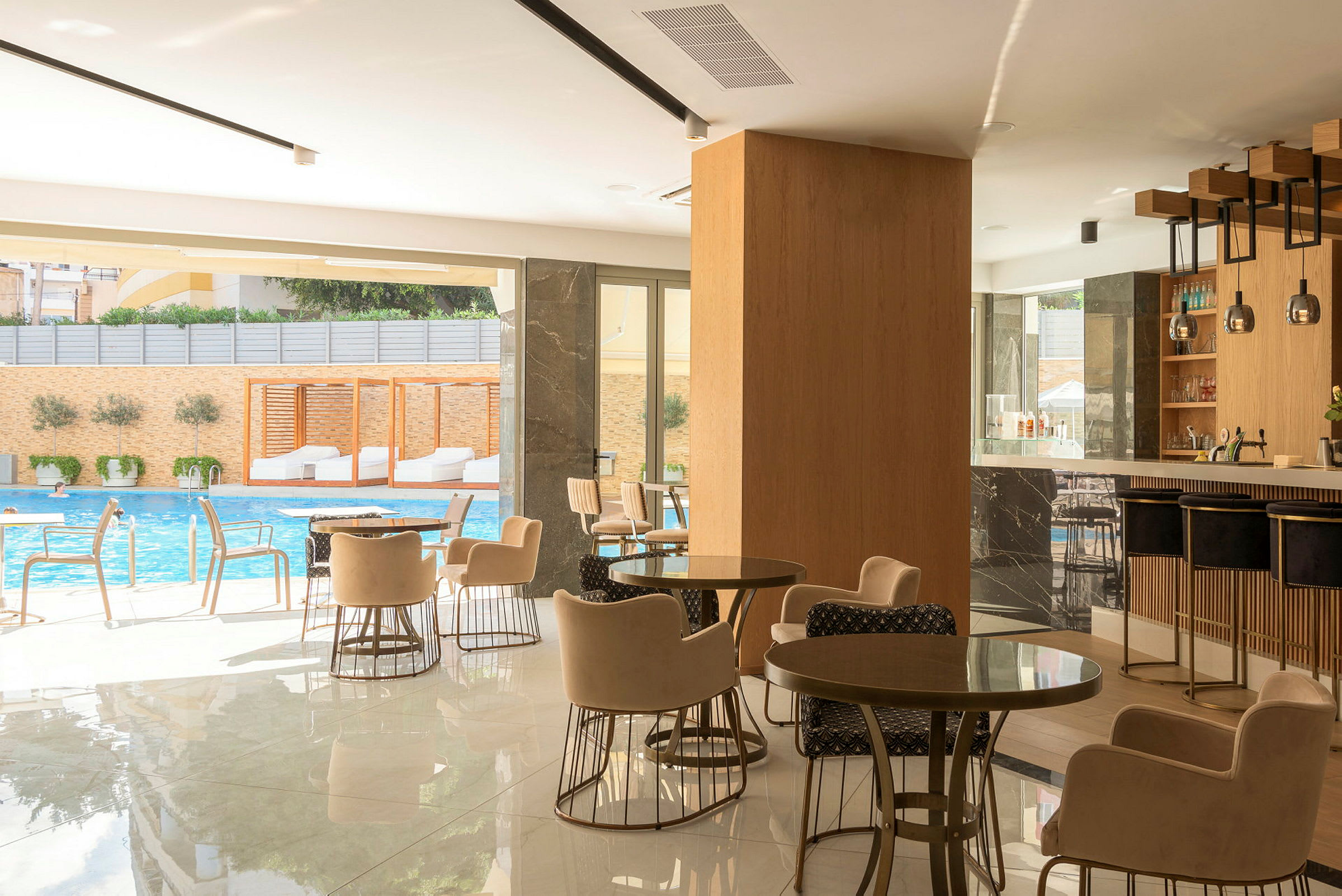 Ett modernt kaféområde med sittgrupper, en bar och utsikt mot en swimmingpool utanför. (AI-genererad text)