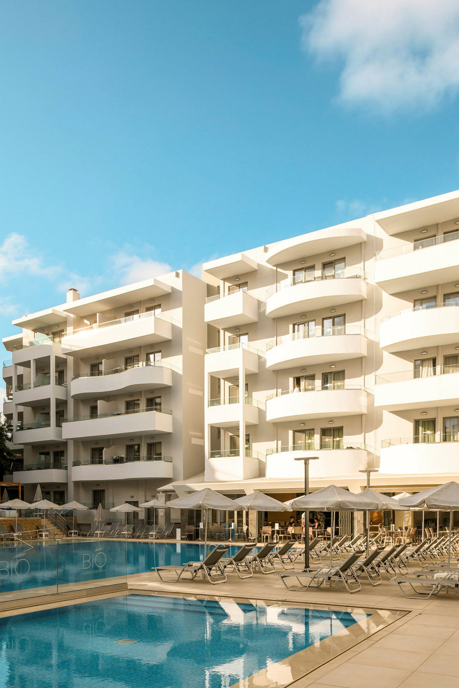 Ett hotell med vita balkonger vid en pool med solstolar och parasoller. (AI-genererad text)