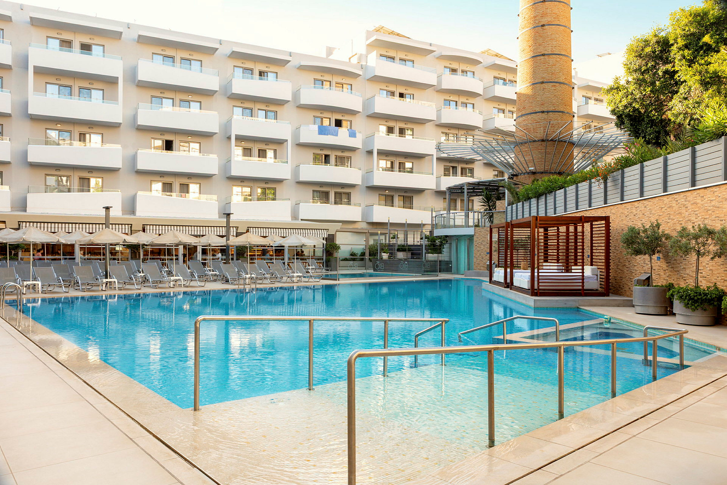 En utomhuspool framför en flerbostadsbyggnad med balkonger. Poolområdet är omringat av solstolar och parasoller. (AI-genererad text)