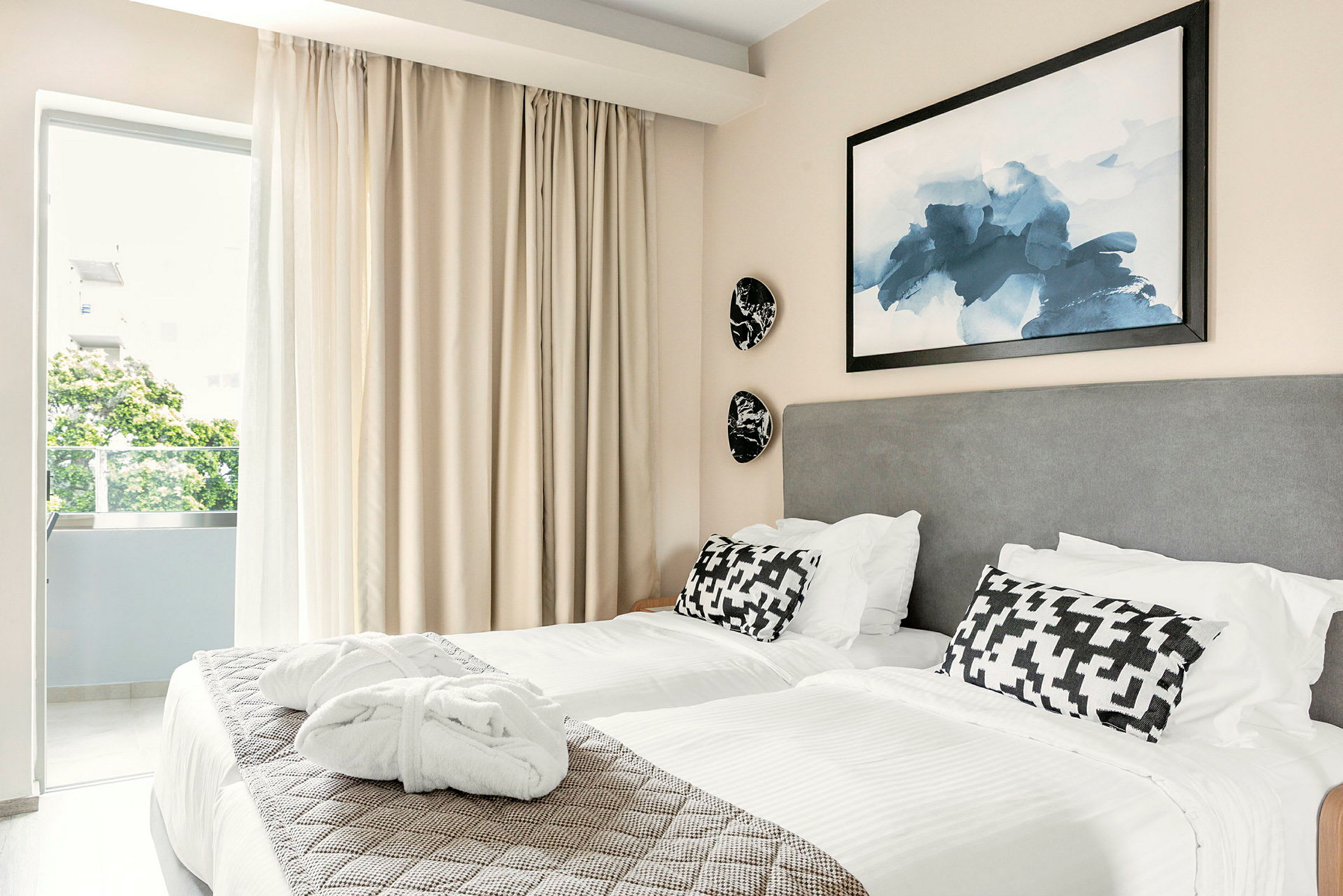 Två sängar med vita lakan och dekorativa kuddar i ett hotellrum. En tavla hänger ovanför sängarna och gardiner är dragna för fönstret. (AI-genererad text)