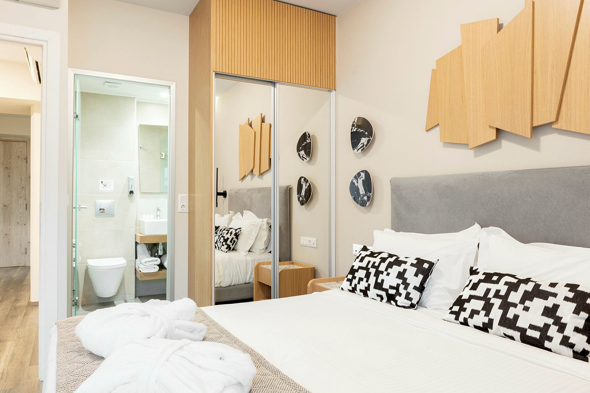Ett hotellrum med en säng, dekorativa kuddar och träkonst på väggen. Till vänster syns ett öppet badrum. (AI-genererad text)