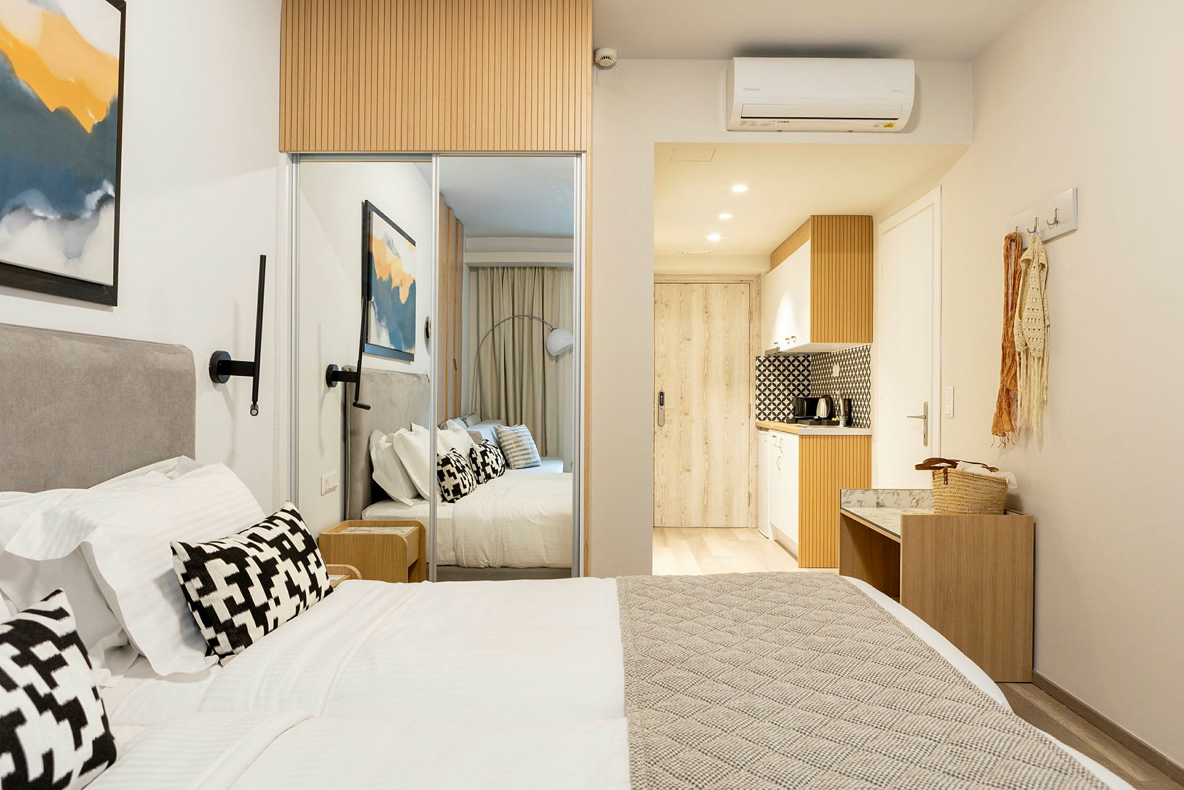 Ett modernt hotellrum med en dubbelsäng, vita sängkläder och dekorativa kuddar. På väggen hänger en abstrakt tavla och i bakgrunden syns ett litet kök. (AI-genererad text)