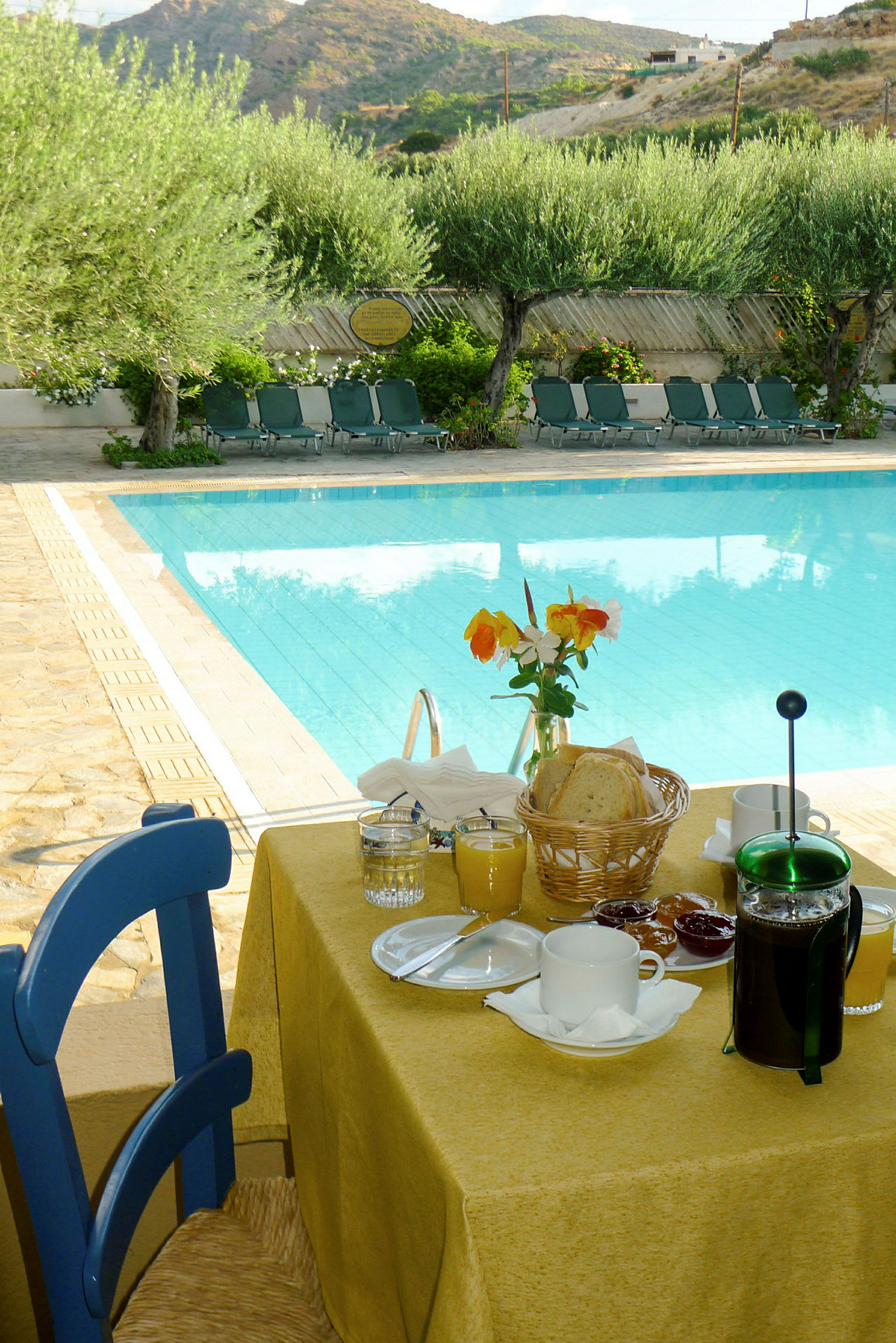 En frukostdukning med utsikt över en pool och solstolar, samt olivträd och berg i bakgrunden. (AI-genererad text)