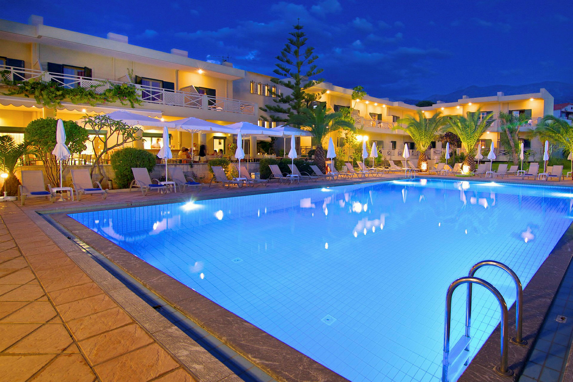 Kvällsbild av ett upplyst poolområde framför ett hotell med solstolar kring poolen. (AI-genererad text)