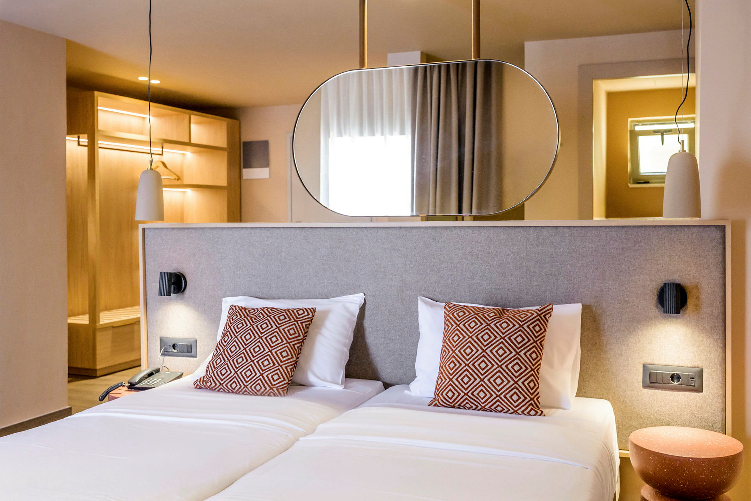 Ett hotellrum med två enkelsängar, dekorativa kuddar och en modern inredning. Bakom sängen finns en spegel och garderob i trä. (AI-genererad text)