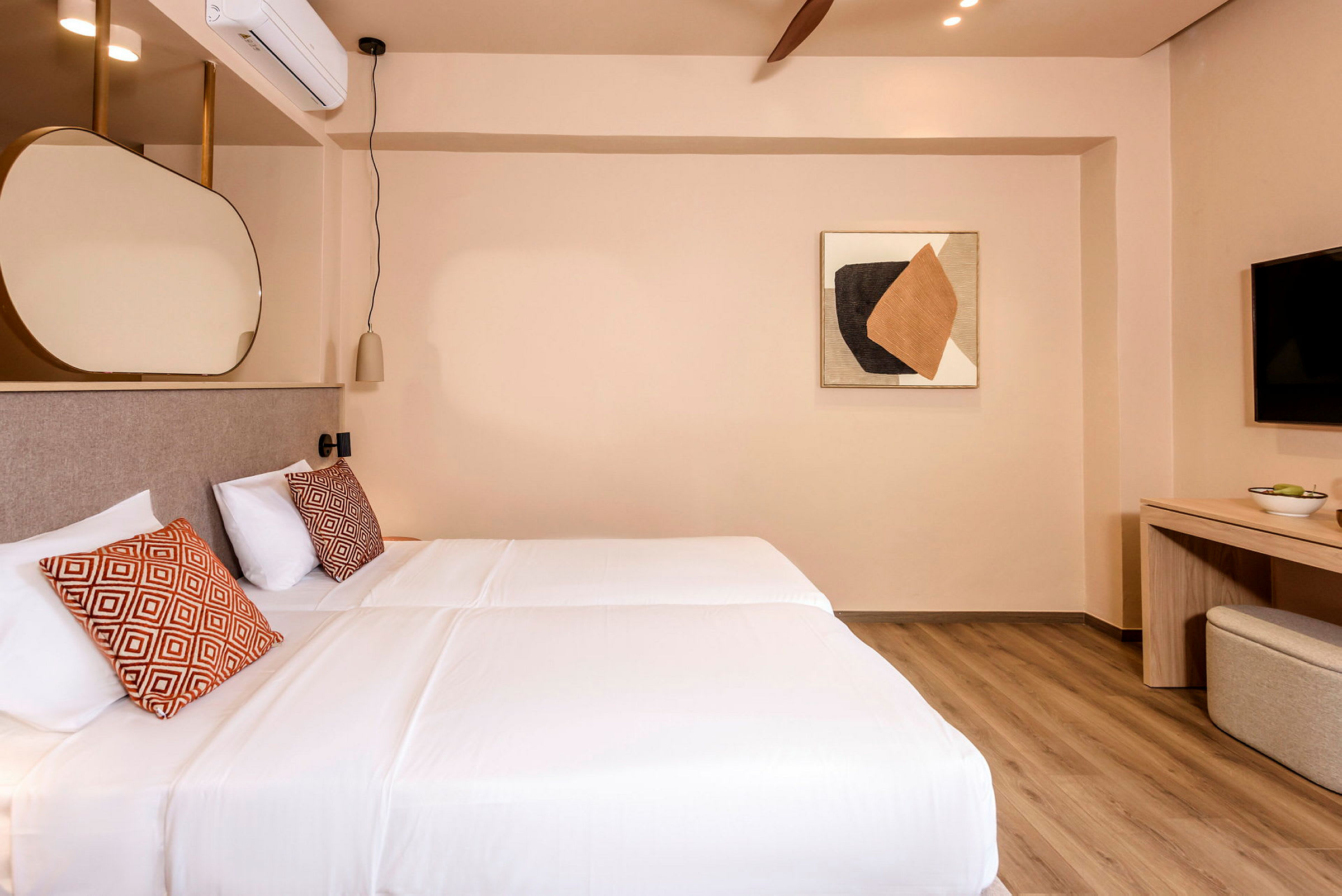 Ett hotellrum med två enkelsängar med vita sängkläder och dekorativa kuddar. Rummet har en väggmonterad TV, en byrå och abstrakt konst på väggen. (AI-genererad text)