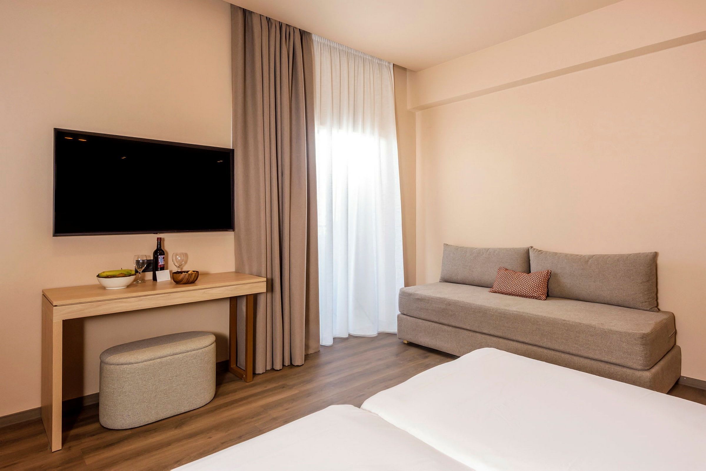 Ett hotellrum med en grå soffa, ett bord med dryck och frukt, en väggmonterad TV och gardiner vid ett fönster. (AI-genererad text)