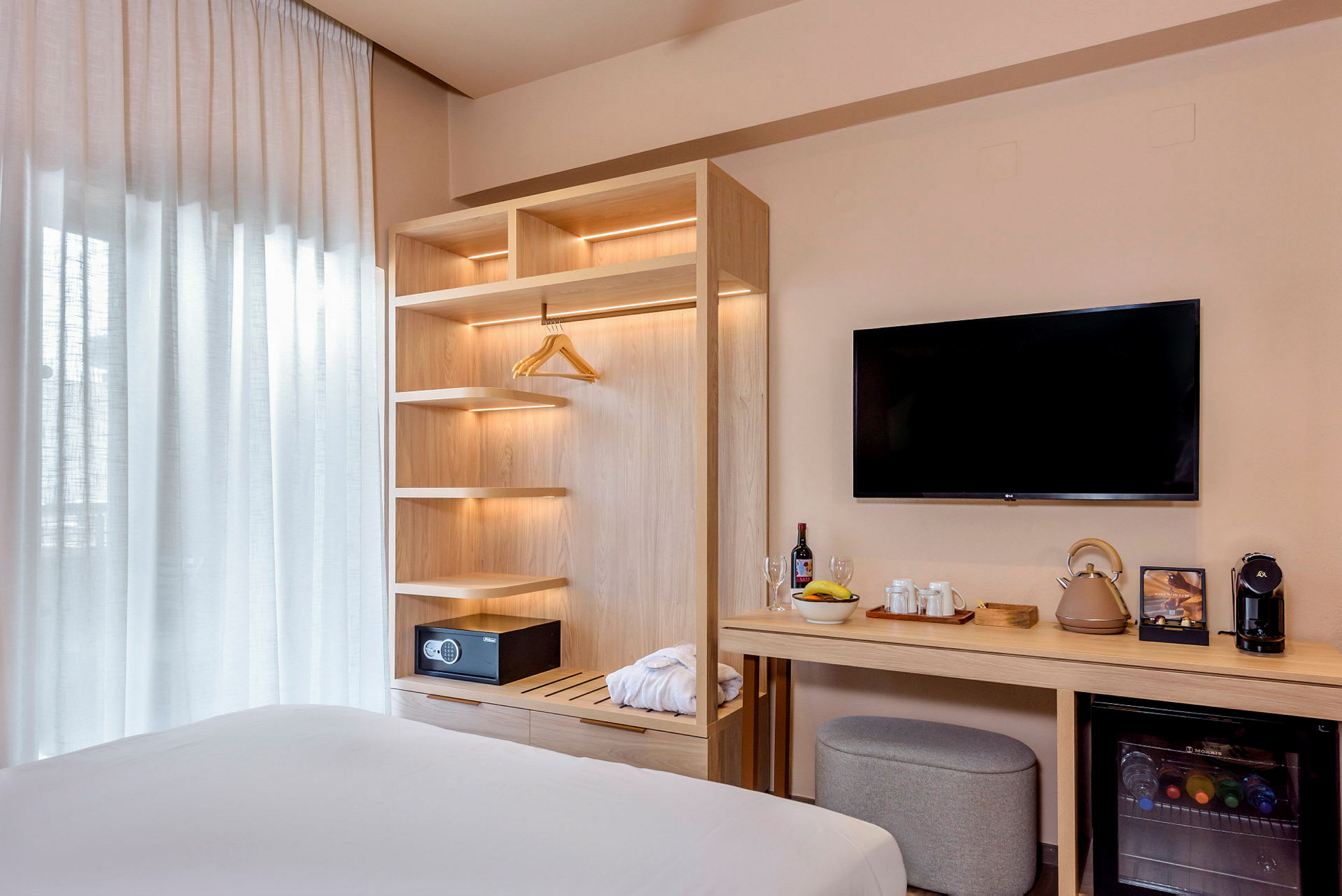 Ett hotellrum med en öppen garderob, en säkerhetsbox, ett bord med drycker och bekvämligheter, samt en väggmonterad TV. (AI-genererad text)