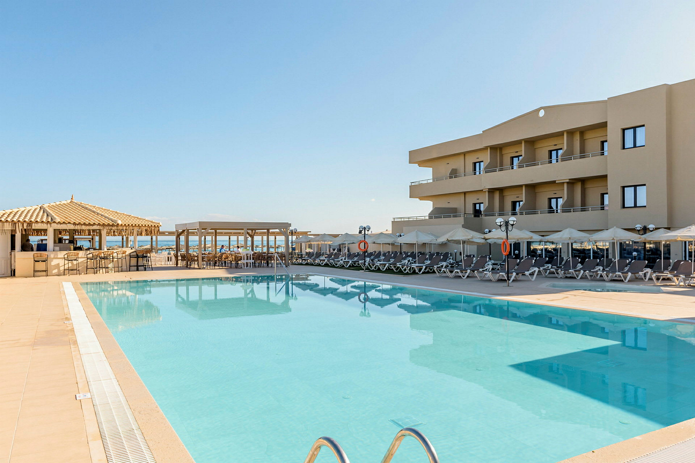 Utomhuspool med solstolar vid ett hotell, med en bar och havet i bakgrunden. (AI-genererad text)