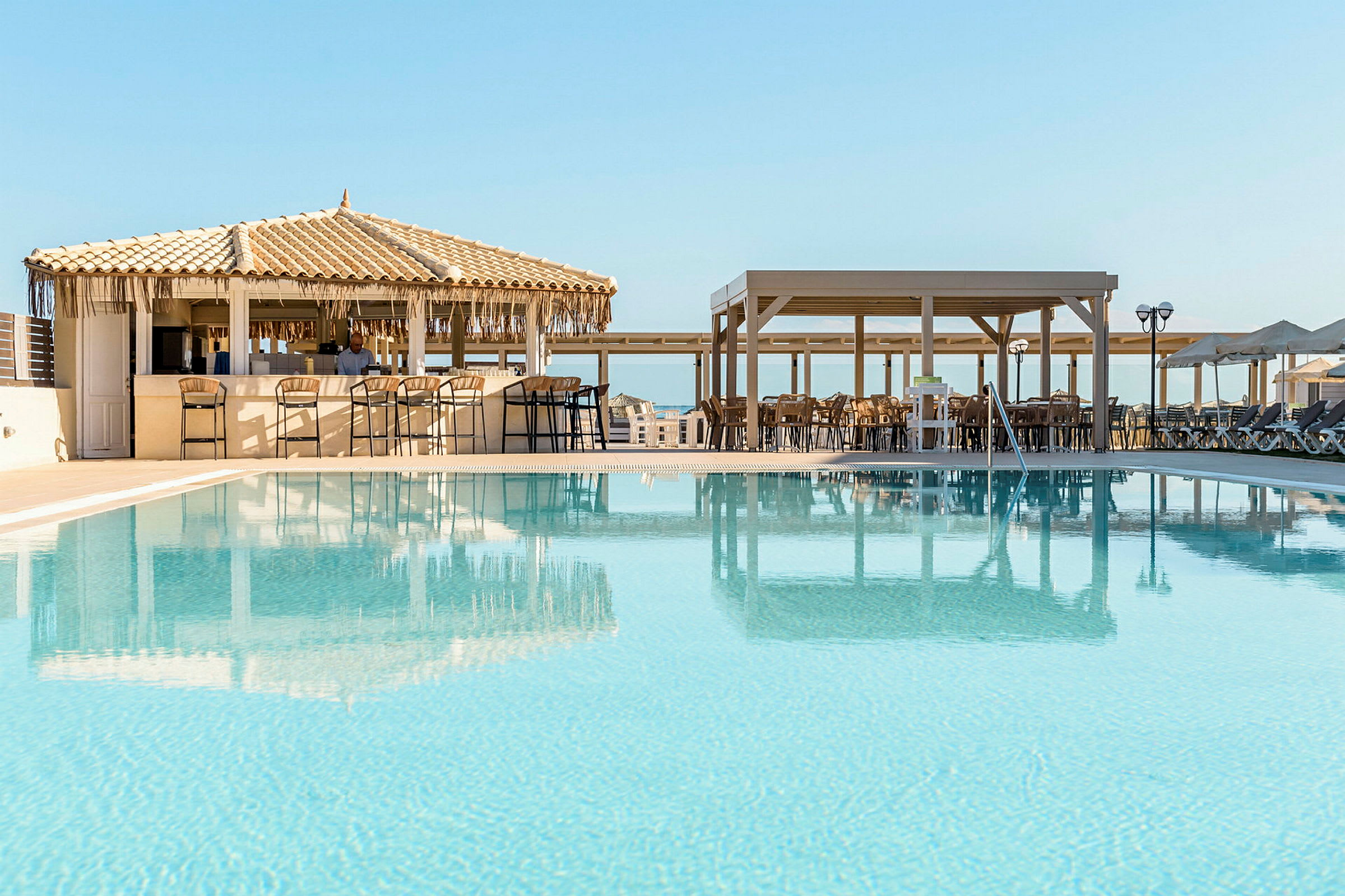En utomhuspool med klarblått vatten och solstolar. I bakgrunden syns en poolbar med halmtak och sittplatser, samt en täckt terrass. (AI-genererad text)