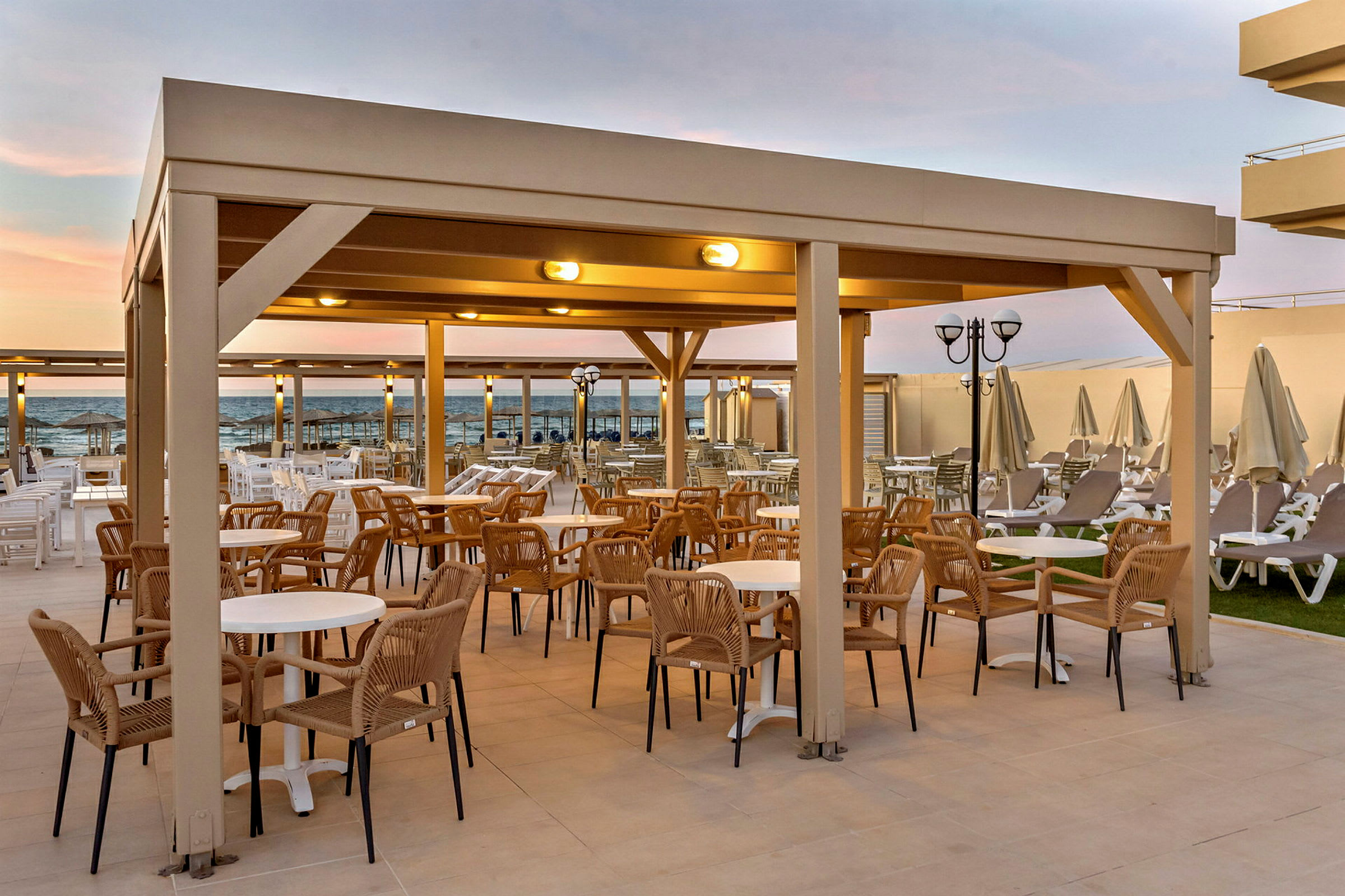 En utomhusterrass med sittplatser och bord under en pergola. Havet syns i bakgrunden. (AI-genererad text)