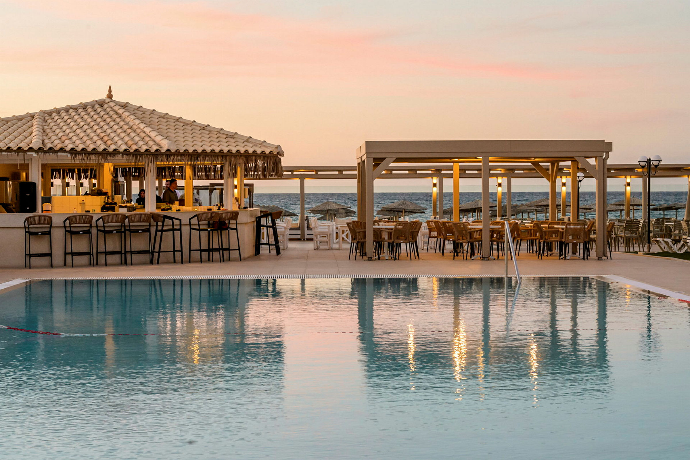 En pool med sittgrupper och en bar i bakgrunden vid havet under en solnedgång. (AI-genererad text)