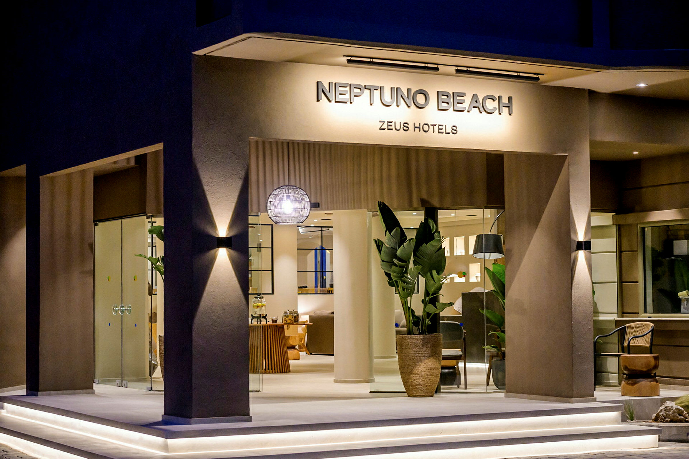 Ingången till hotell Neptuno Beach med glasdörrar, växtarrangemang och modern belysning. (AI-genererad text)