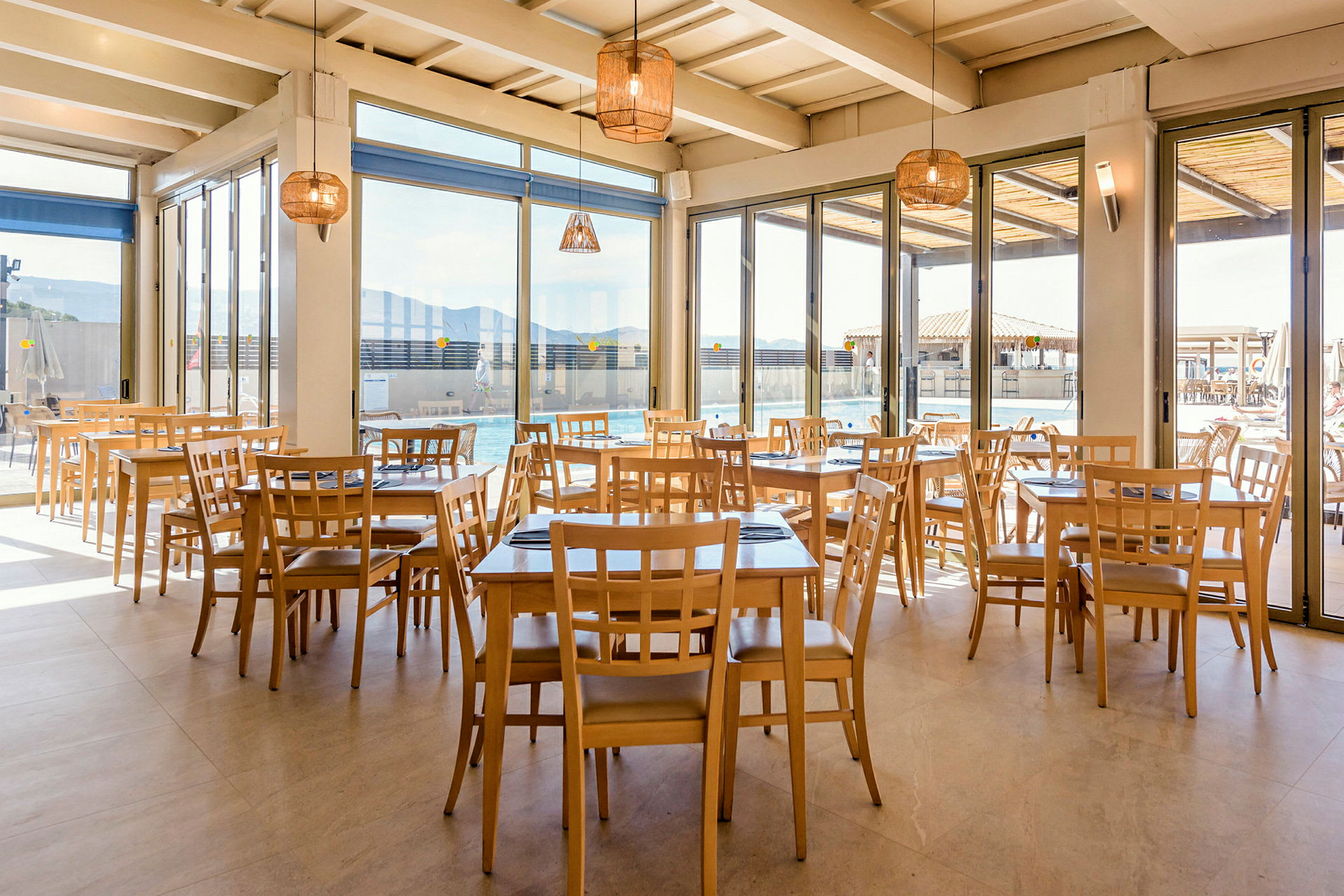 Restaurang med ljusa träbord och stolar, stora fönster mot en utomhuspoolsområde. (AI-genererad text)