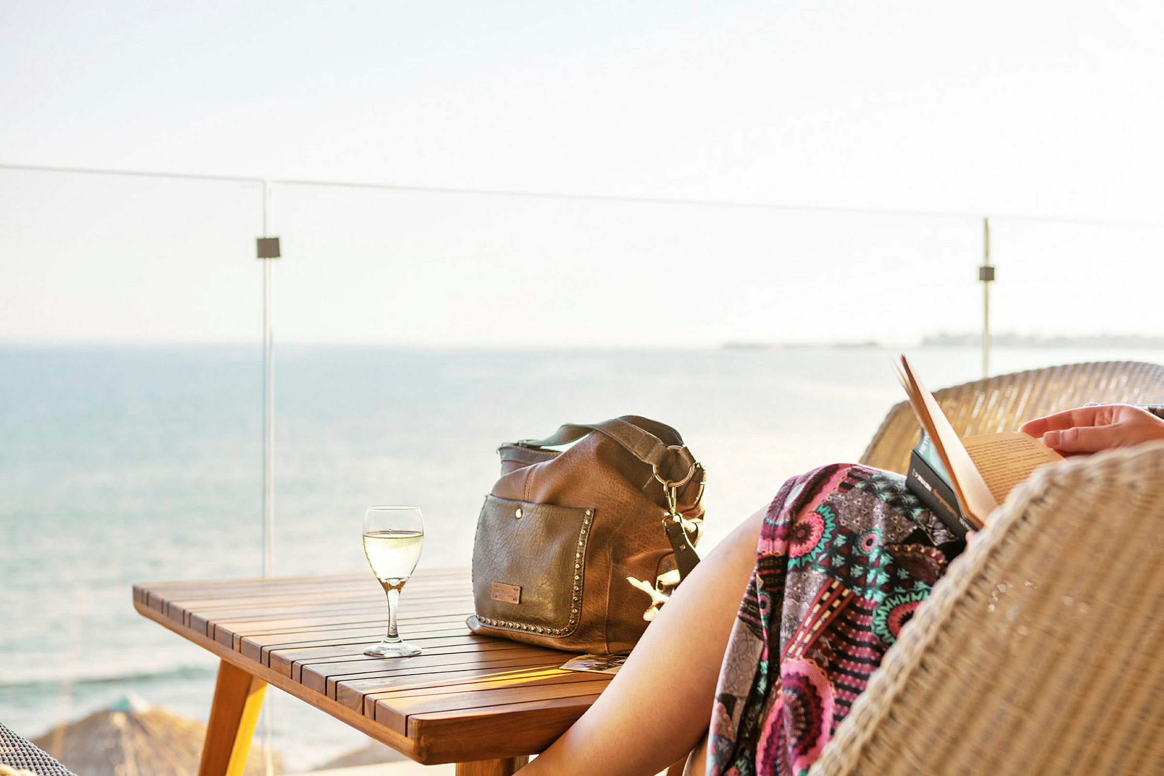 Person sitter och läser en bok vid ett bord med ett glas vin och en handväska, med havet i bakgrunden. (AI-genererad text)