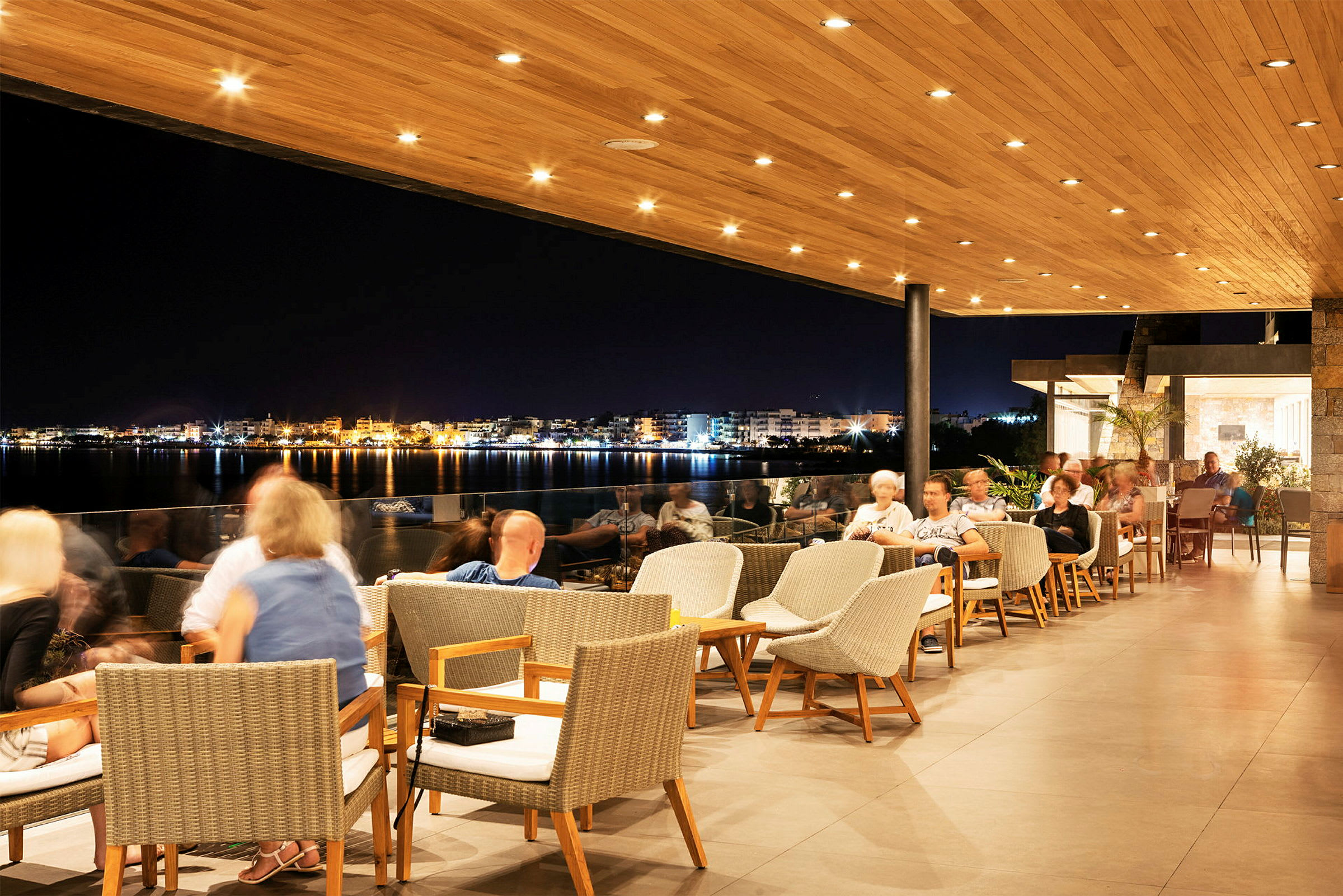 En restaurang med utomhusområde under en träklädd takterrass, med utsikt över en upplyst stad vid vattnet på kvällen. (AI-genererad text)