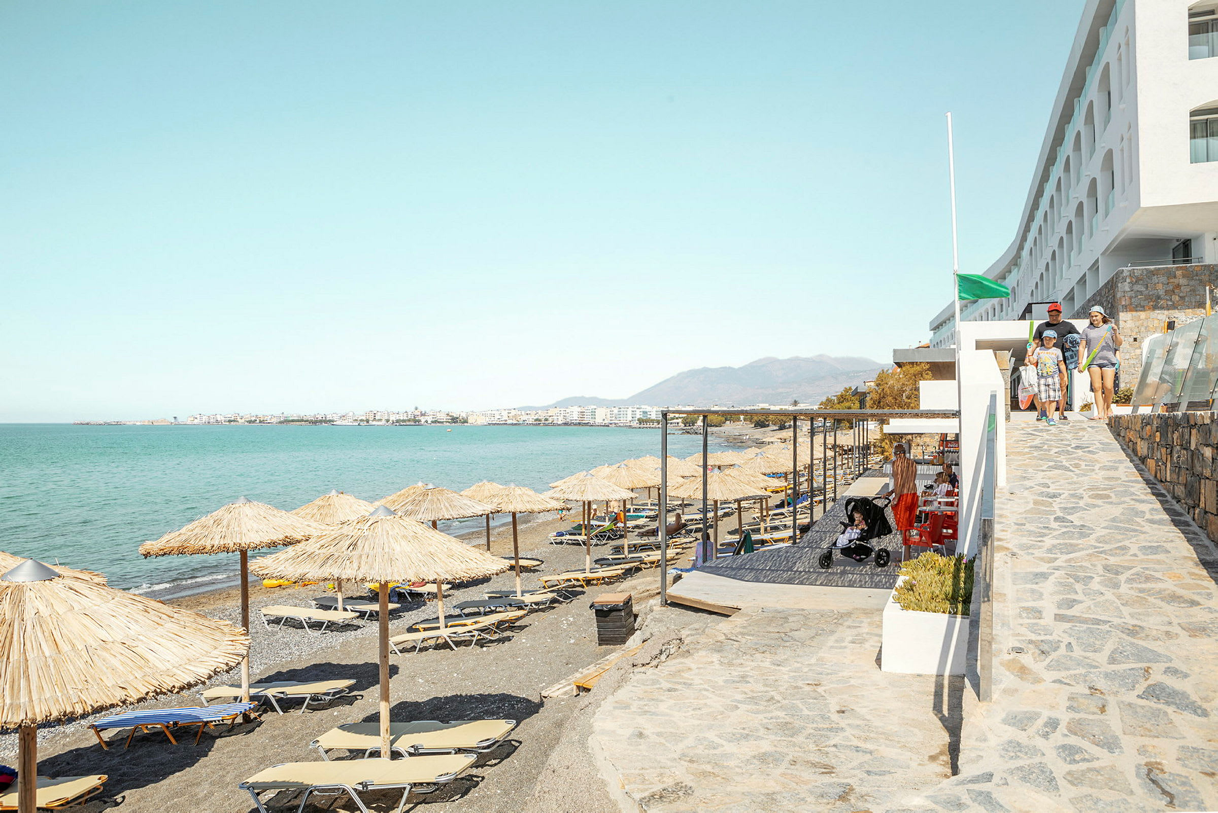 Strand med parasoller och solstolar, havet i bakgrunden och en byggnad till höger. (AI-genererad text)