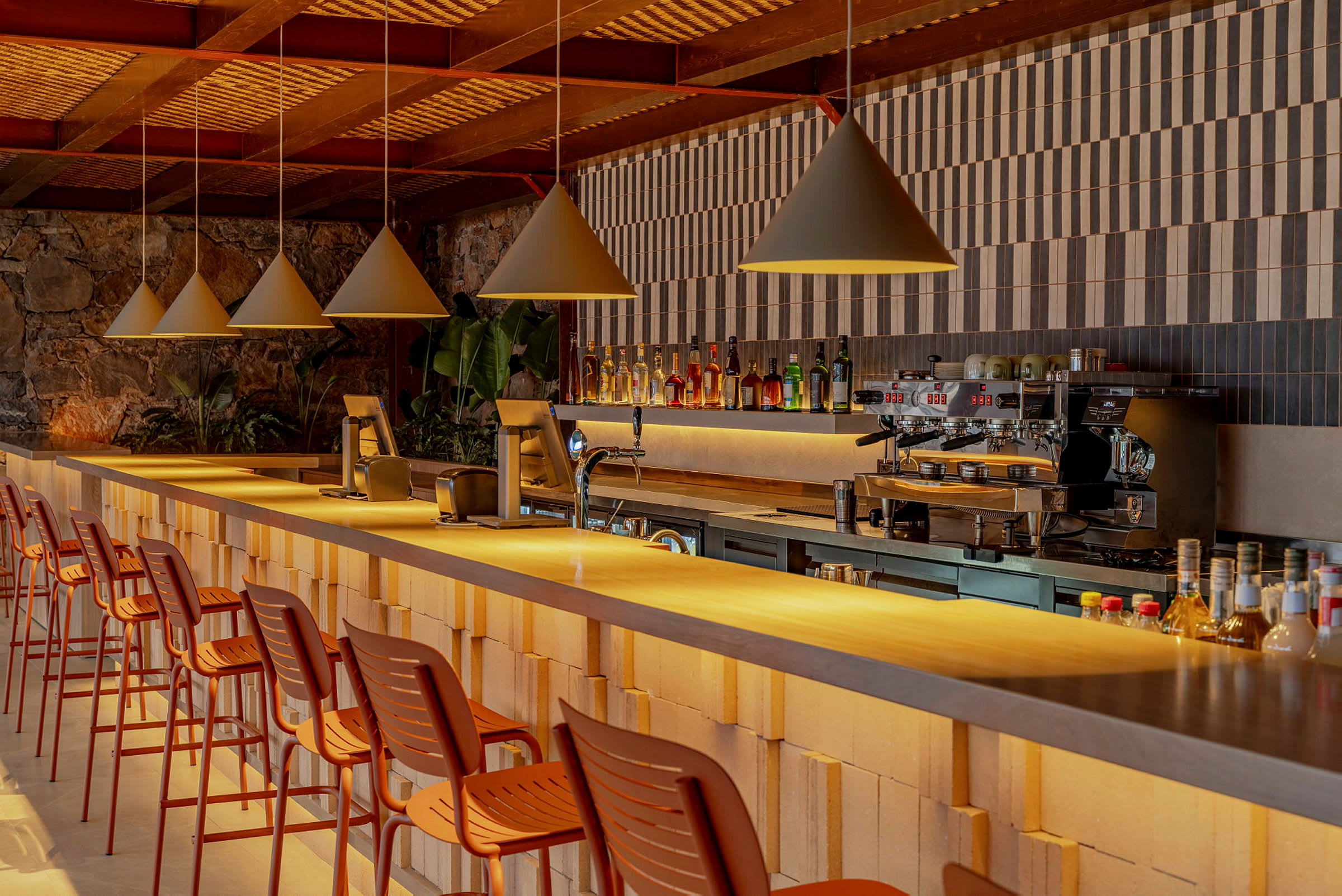 Bilden visar en modern bar med en rad höga stolar vid en lång bardisk. Baren är dekorerad med flaskor och har en kaffemaskin. (AI-genererad text)