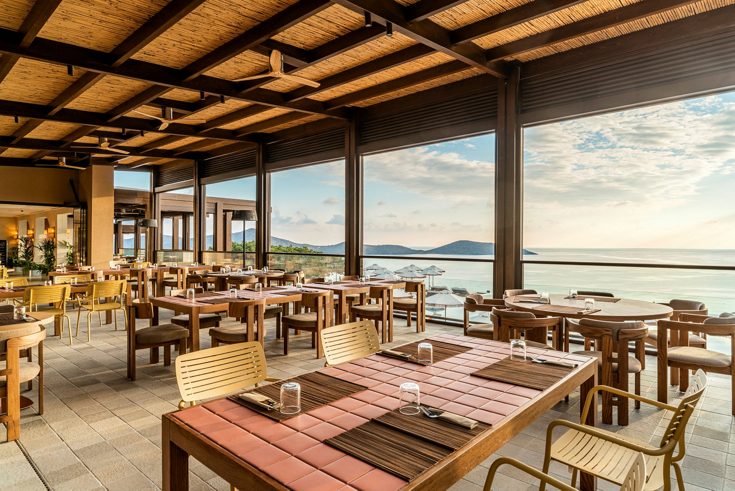 En öppen och ljus restaurang med träbord och stolar, med utsikt över havet och horisonten. (AI-genererad text)