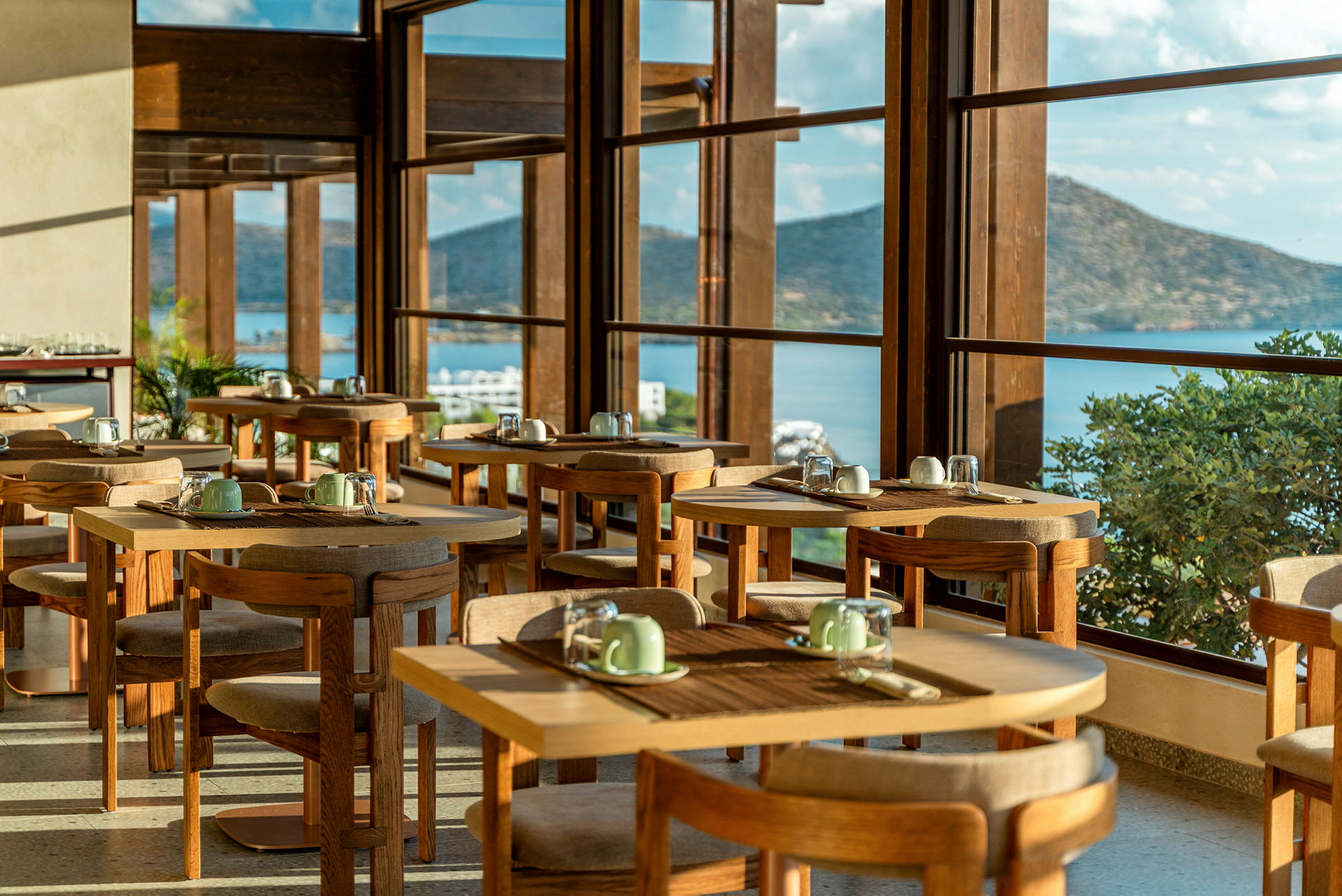 Ett restaurangområde med bord och stolar, med stora fönster som visar en utsikt över en havsvik och berg. (AI-genererad text)
