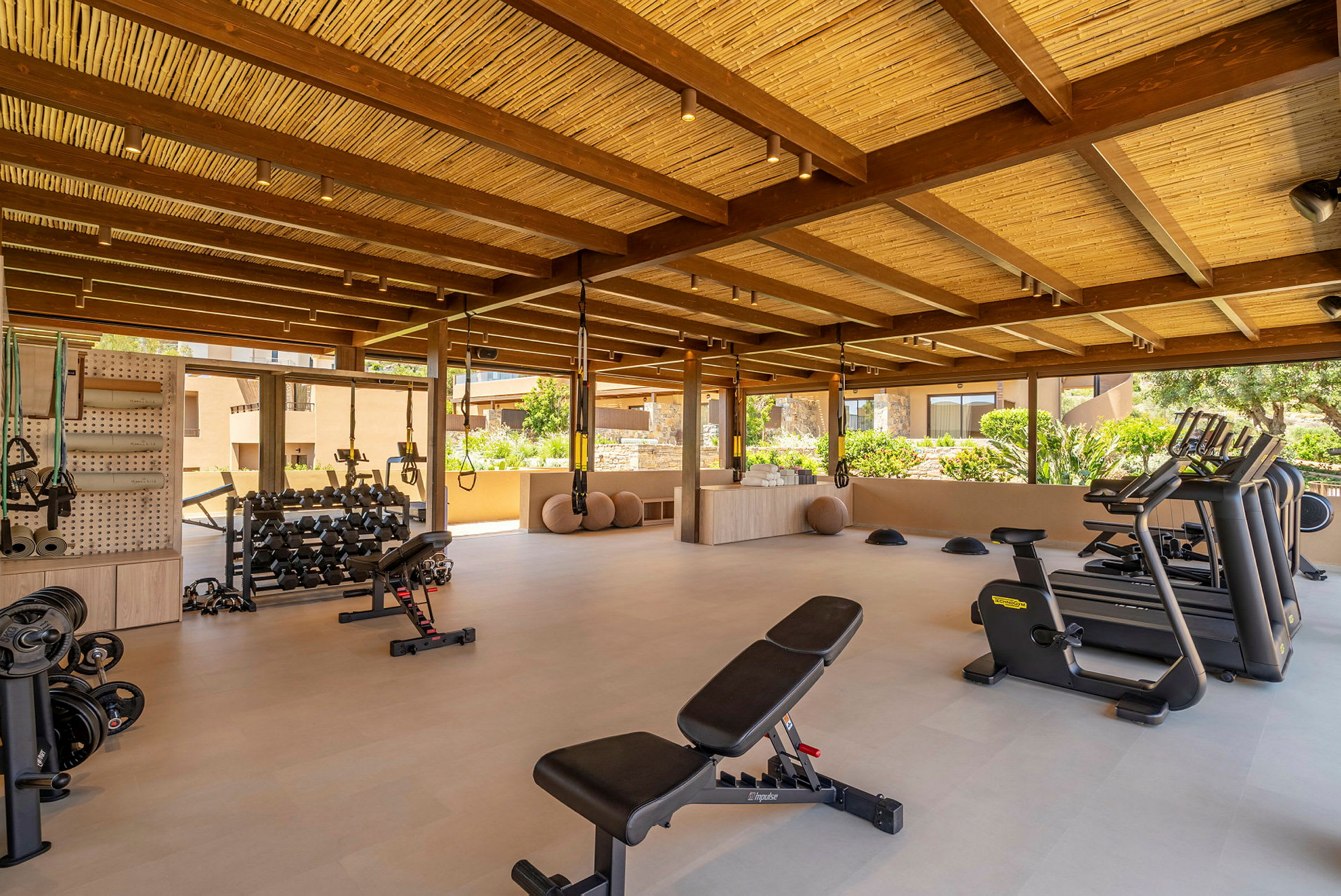 Ett gym med träningsutrustning såsom bänkar, vikter, löpband och cyklar, beläget under ett tak med träbjälkar. (AI-genererad text)