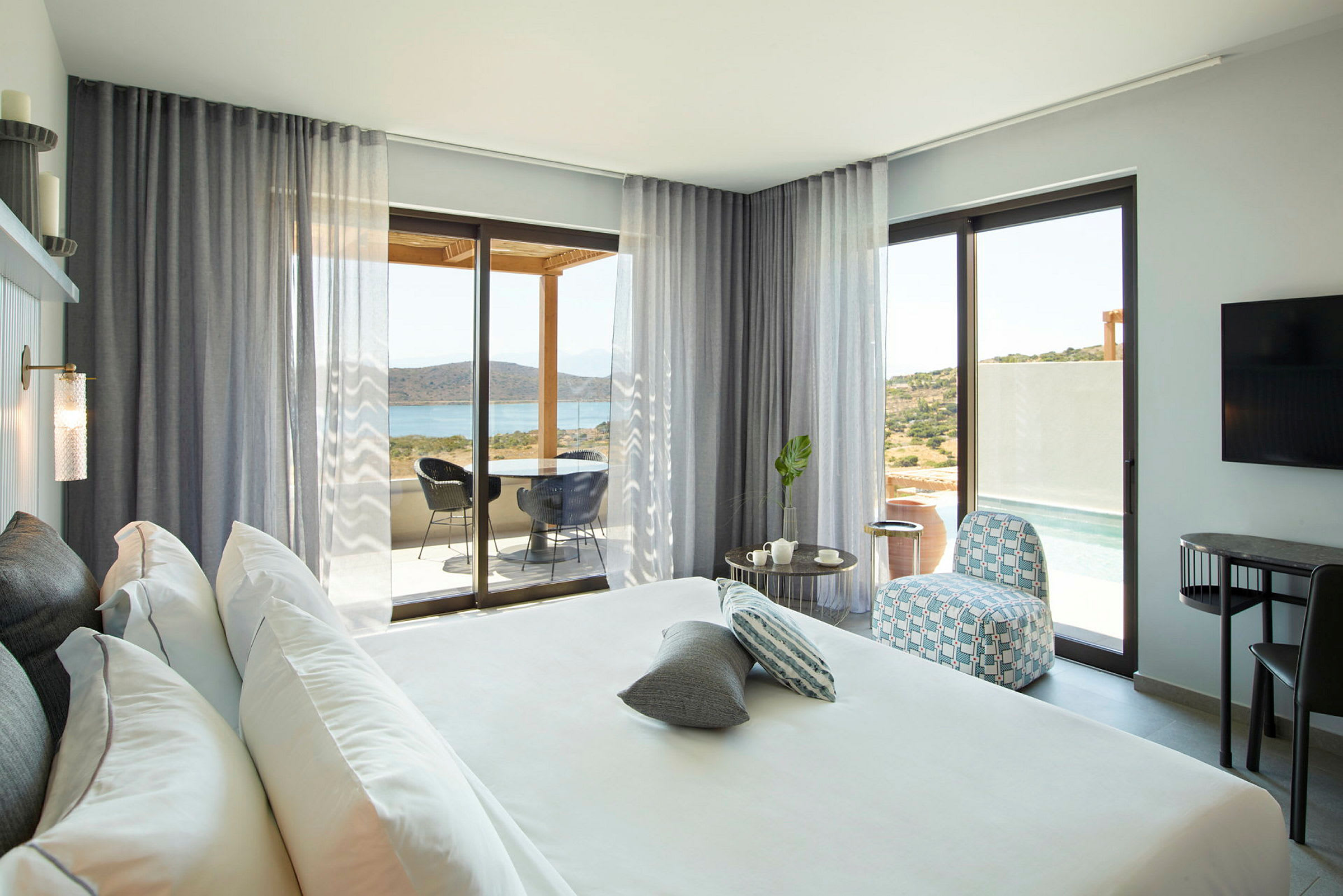Modernt hotellrum med stor säng, sittområde och utsikt mot hav och landskap genom glasdörrar. (AI-genererad text)