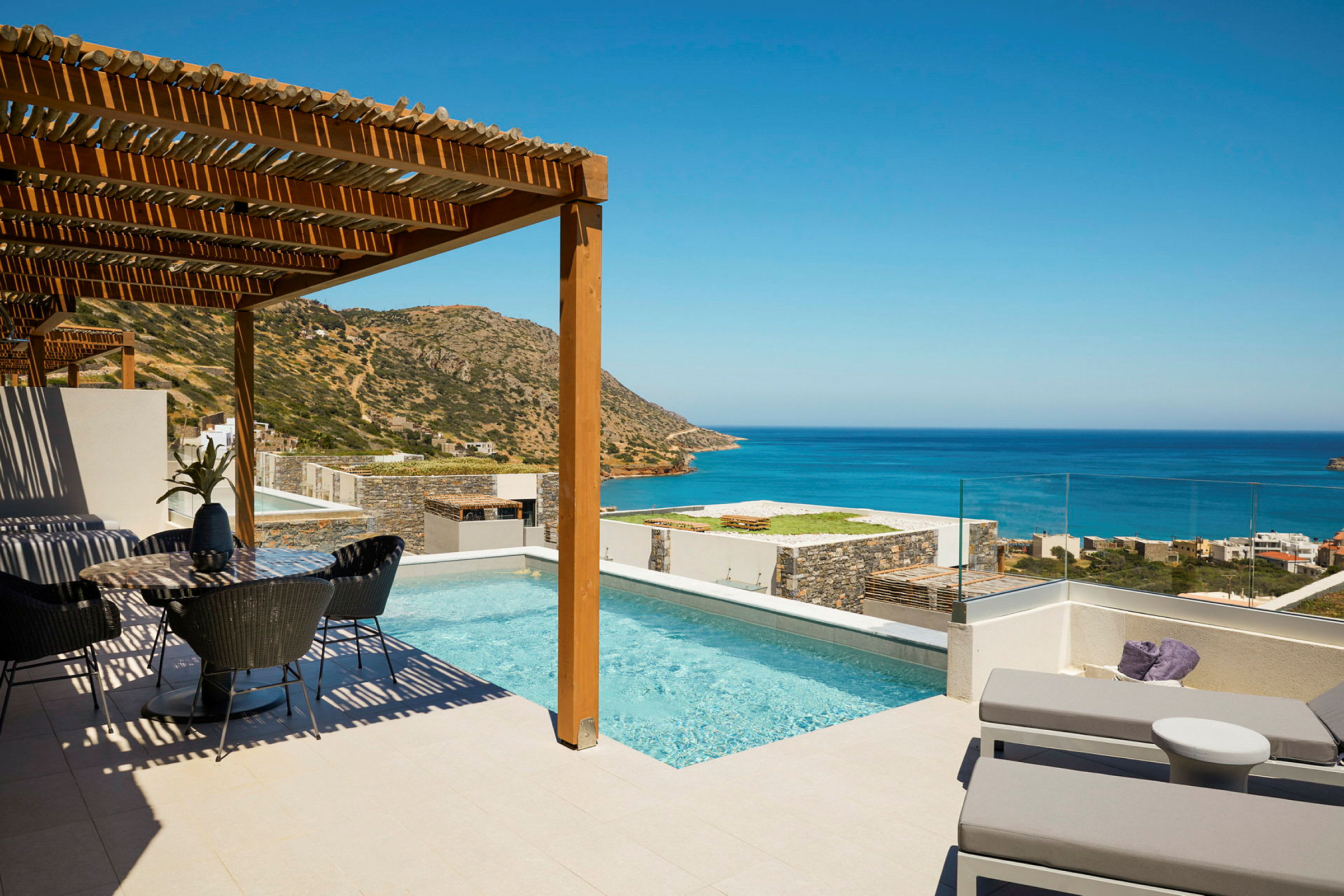 Bild av en terrass med en pool, solsängar och en matplats, med utsikt över havet och berg under en klar himmel. (AI-genererad text)
