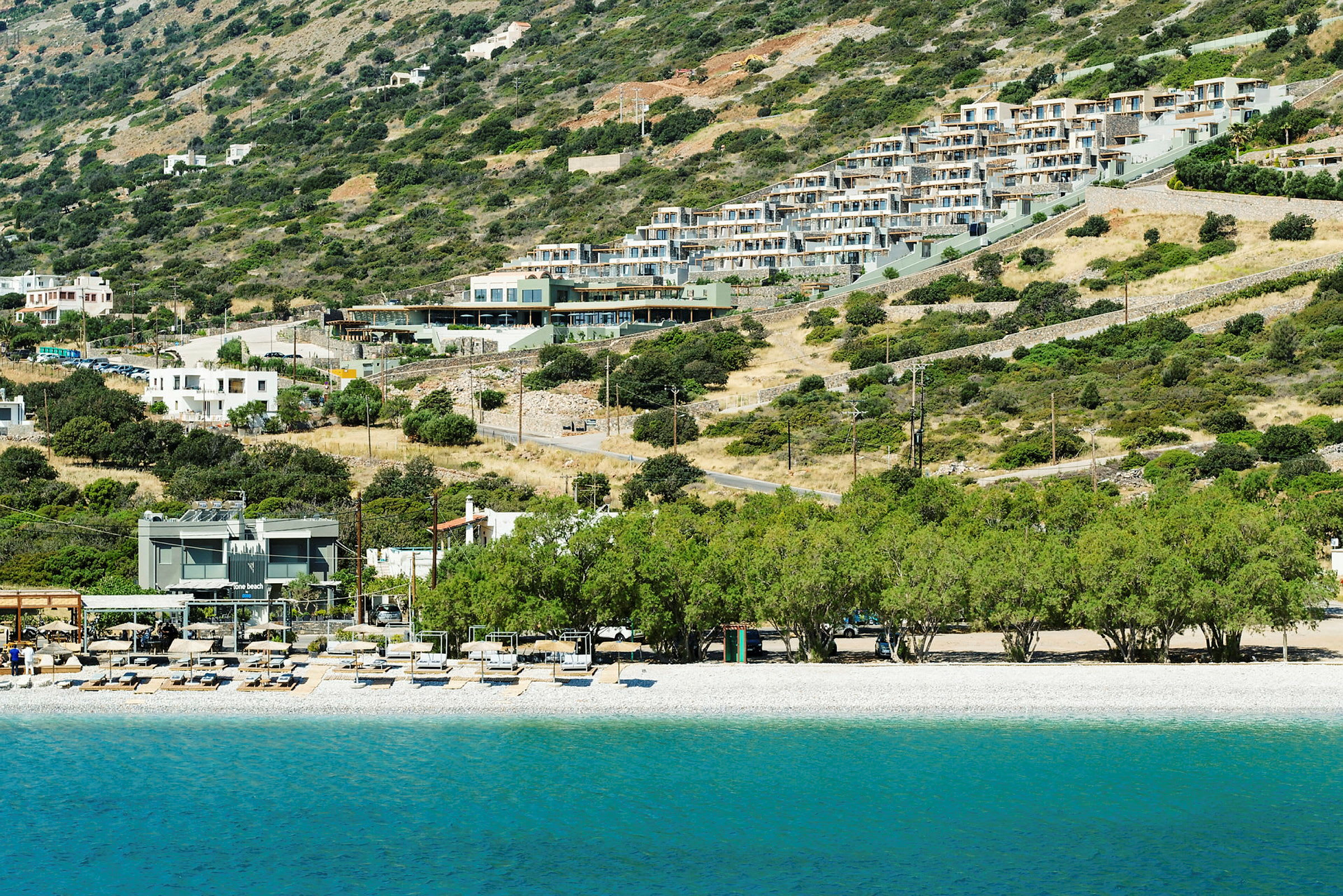 Bild av en semesterort belägen på en sluttning med byggnader och vegetation ovanför en strand. (AI-genererad text)