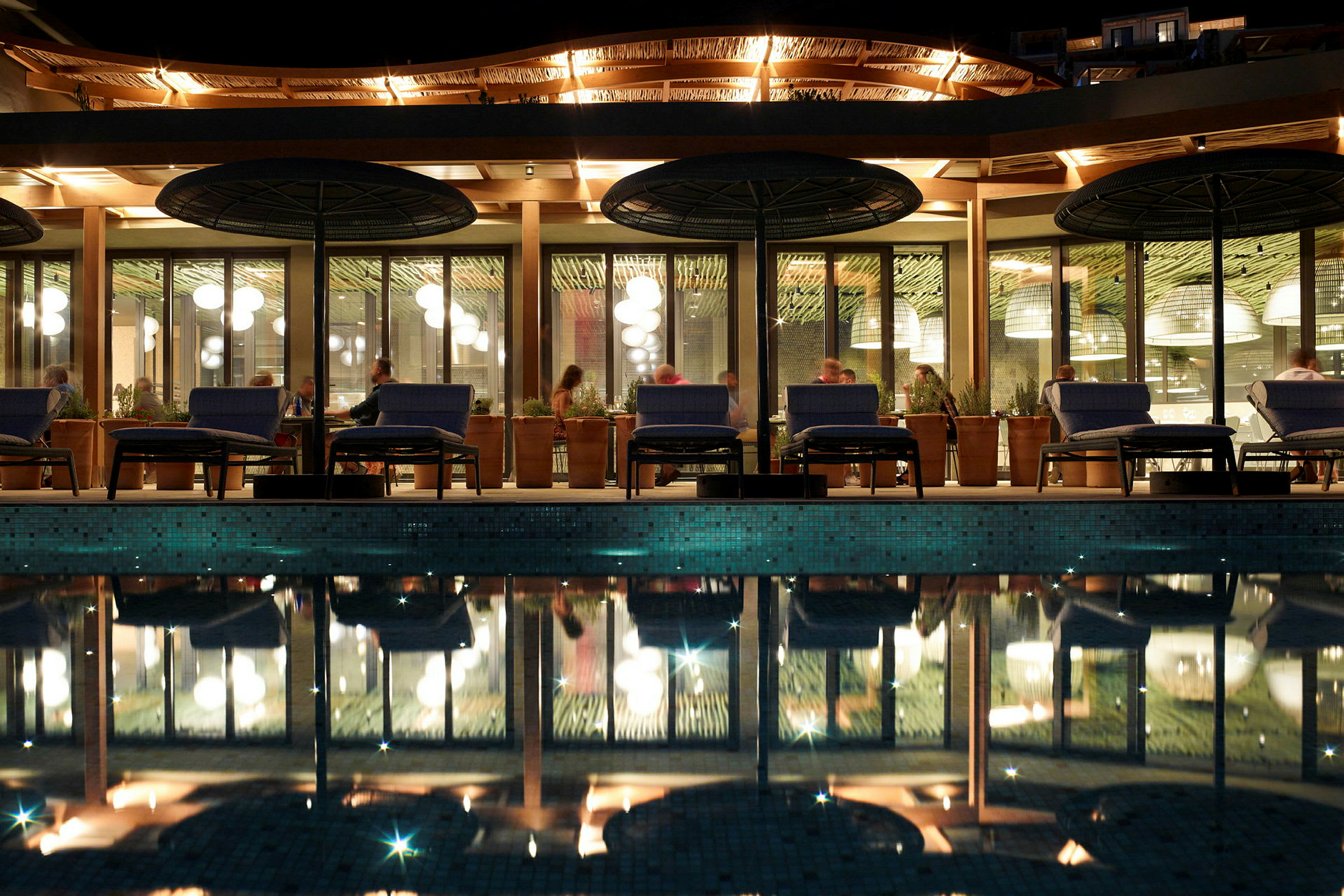 Bild av utomhusområdet vid en restaurang med kvällsbelysning, en pool i förgrunden och gäster som sitter vid bord. (AI-genererad text)