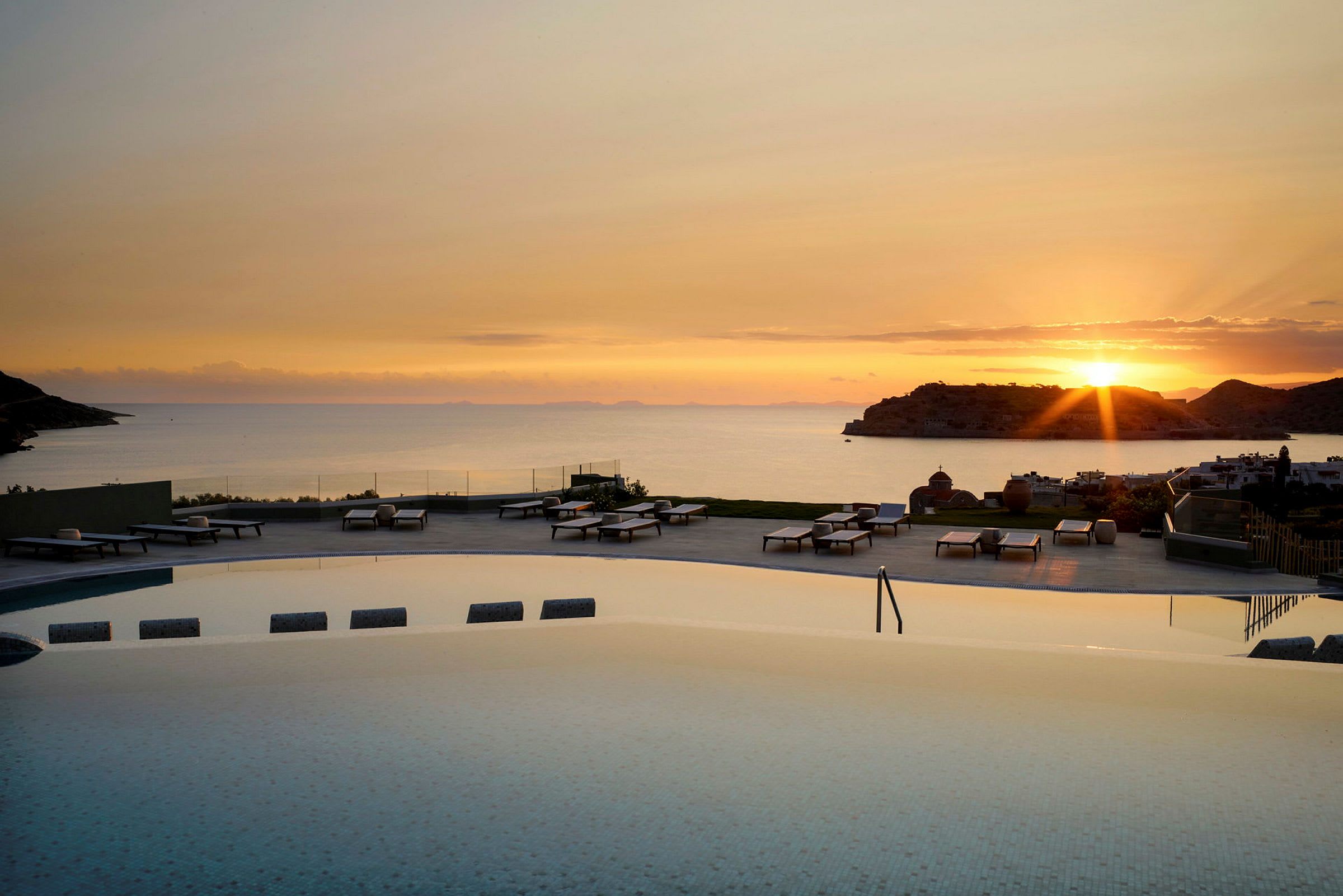 Utsikt över en pool med solstolar i förgrunden och havet vid solnedgång i bakgrunden. (AI-genererad text)