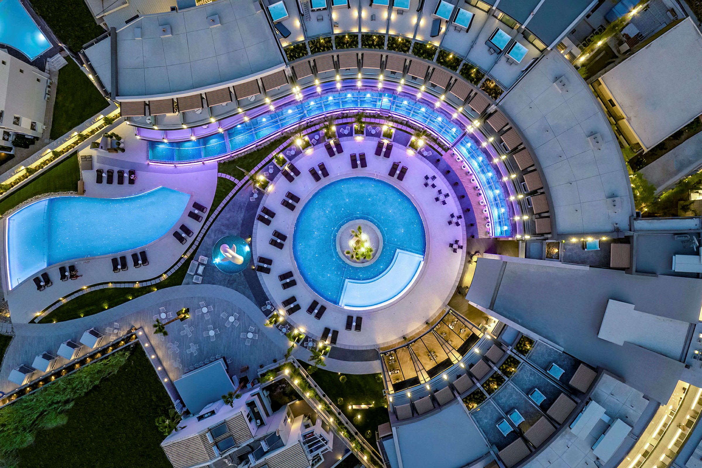 Flygbild över ett hotellkomplex med en rund pool omgiven av solstolar och byggnader. Belysning skapar en lysande atmosfär. (AI-genererad text)