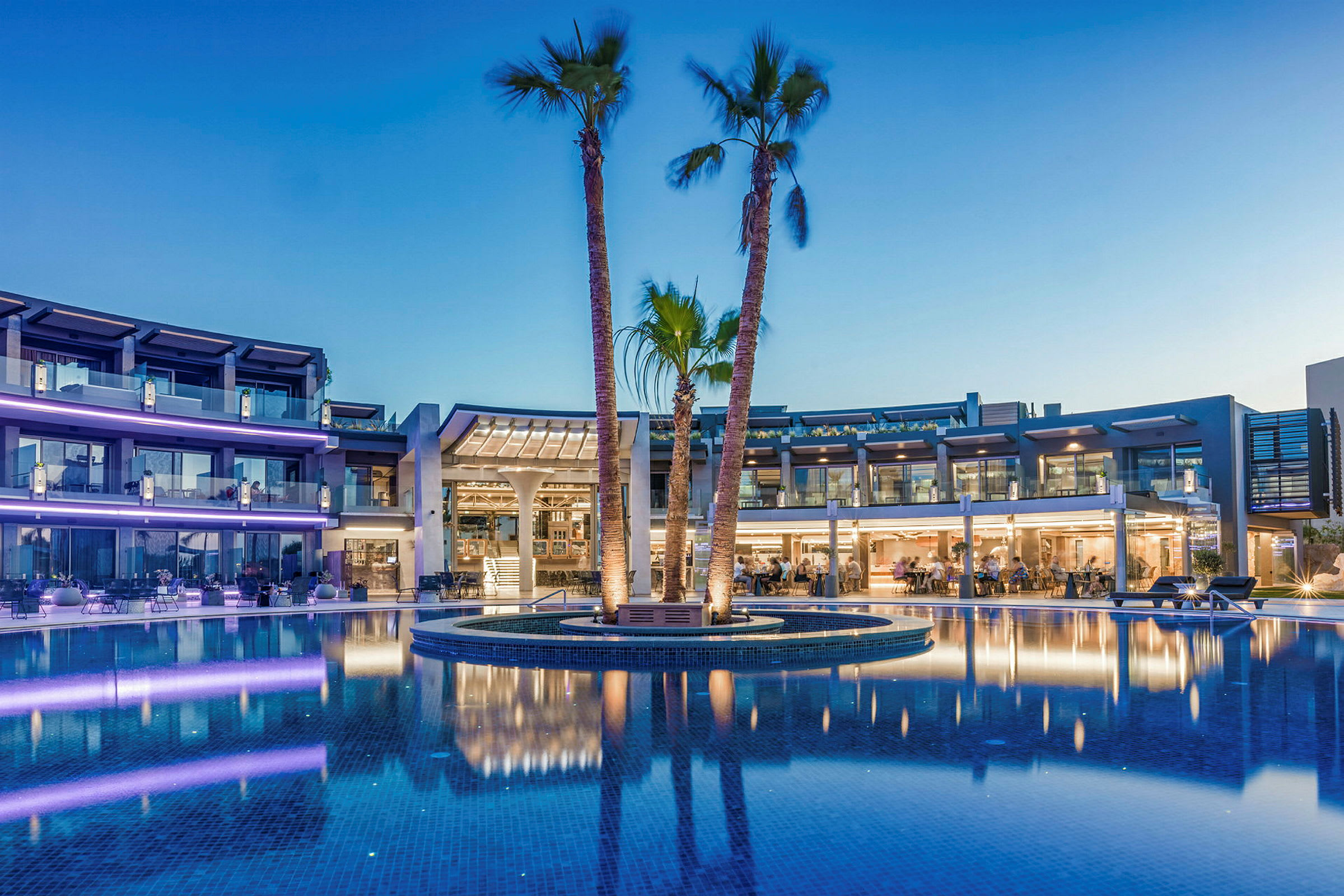Hotellkomplex vid en pool med palmer och belysning i skymning. (AI-genererad text)