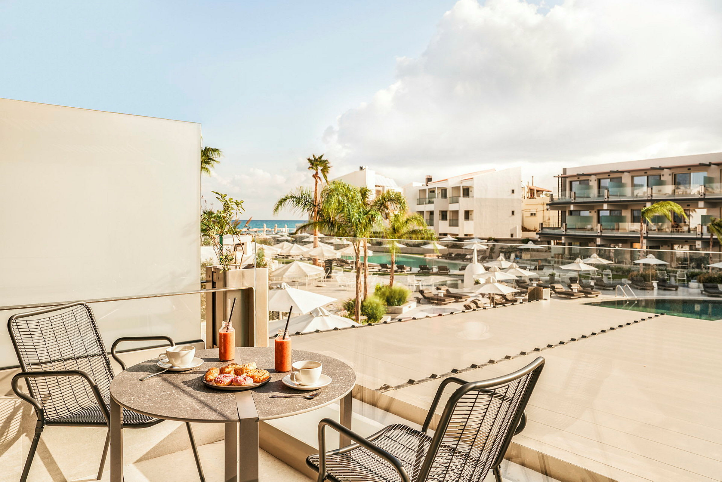 En balkong med bord, stolar och frukost serverad ovanför ett hotellområde med pool och utsikt mot havet. (AI-genererad text)