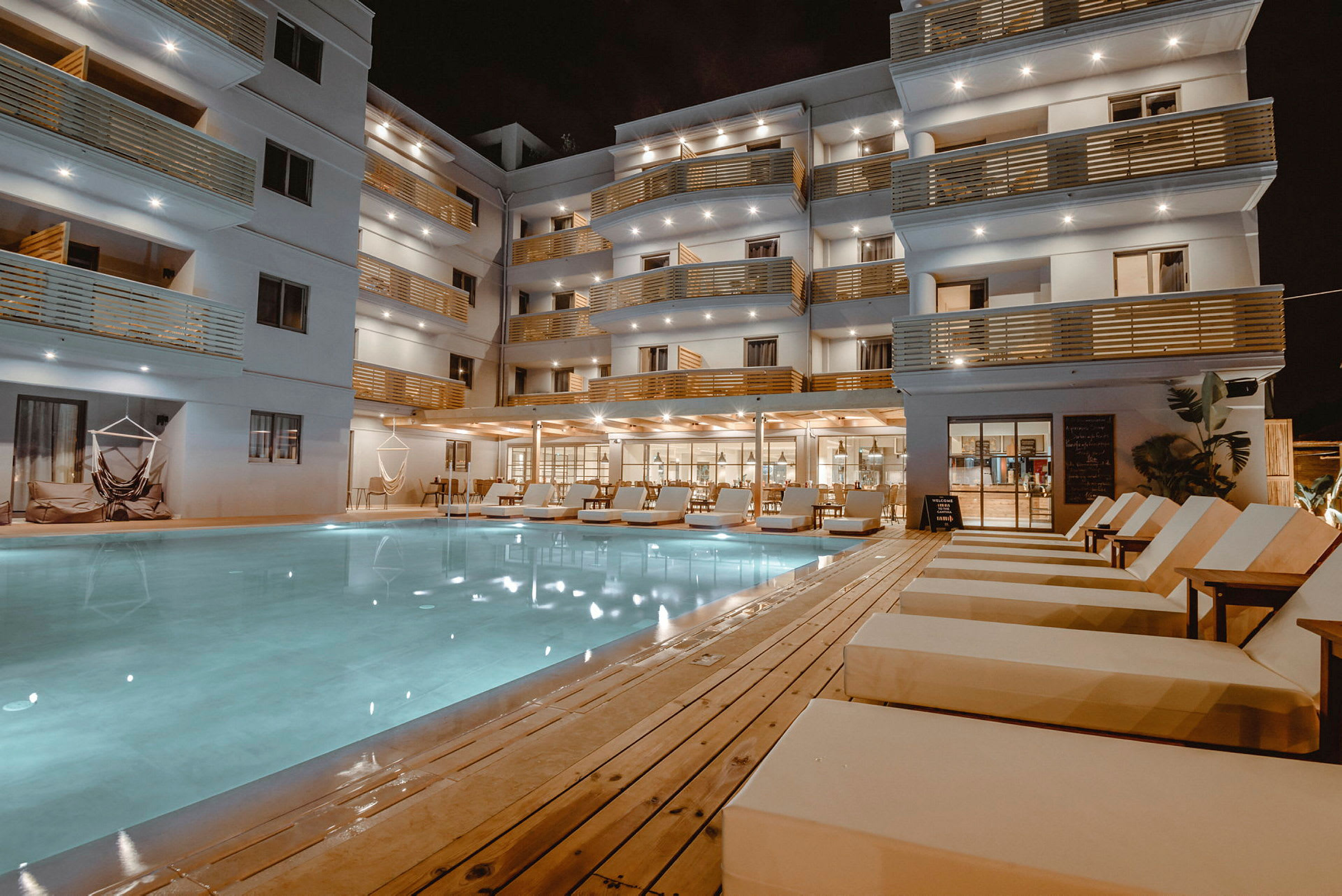 Hotellbyggnad med balkonger och utomhuspool med solstolar. (AI-genererad text)