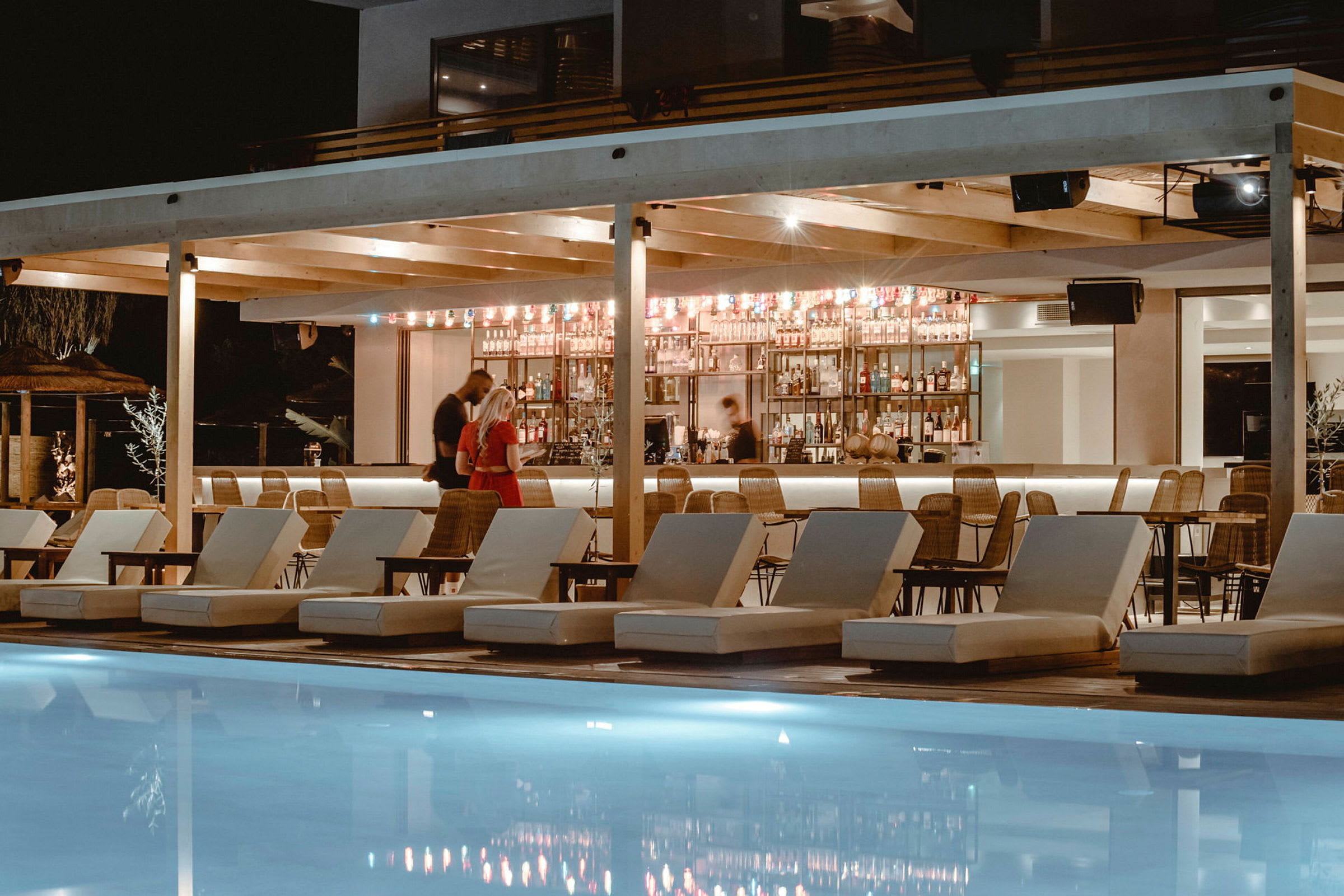 En kvällsmiljö vid en lyxig pool med solstolar framför en upplyst bar. (AI-genererad text)