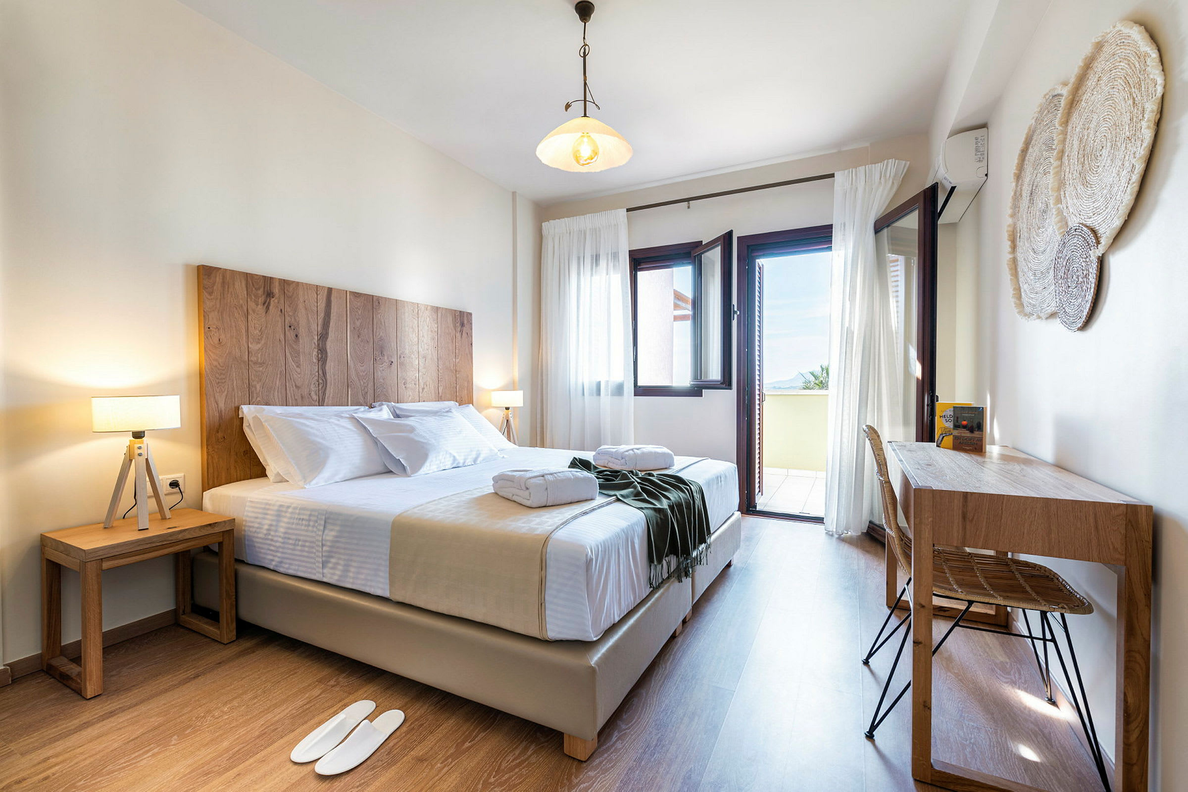 Ett ljust och inbjudande hotellrum med en dubbelsäng, två nattduksbord och ett skrivbord. Fönstren står öppna mot en balkong med utsikt. (AI-genererad text)