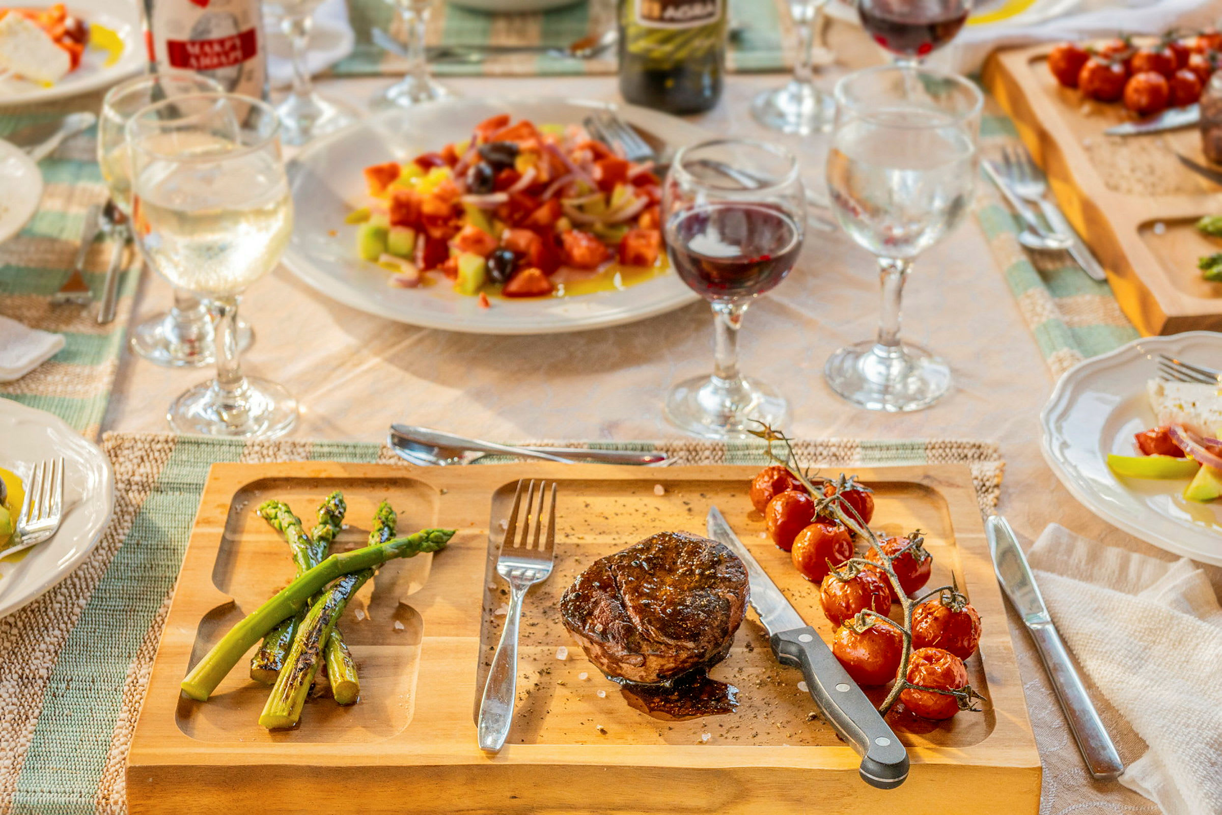 En träbricka med en portion grillat kött, sparris och cocktailtomater, omgiven av dukat bord med glas och tallrikar. (AI-genererad text)