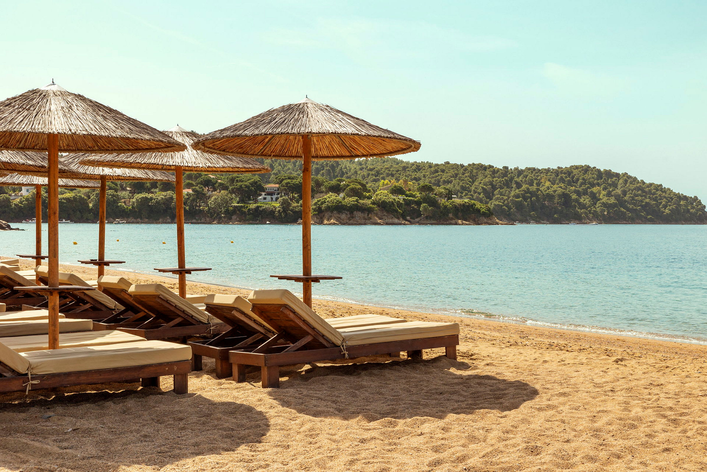 Strand med solstolar och parasoller, hav och skog i bakgrunden. (AI-genererad text)