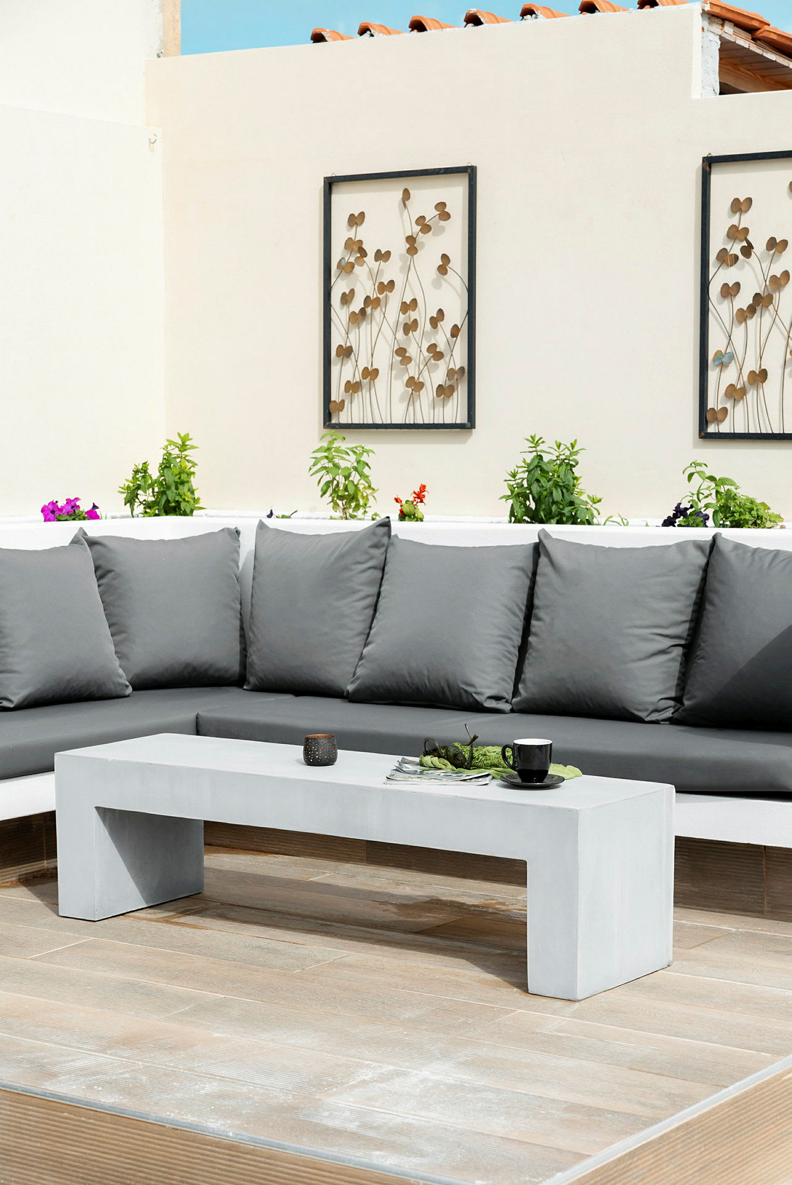 Grå utomhussoffa med kuddar bredvid en vit betongbord omgiven av växter på en terrass. (AI-genererad text)