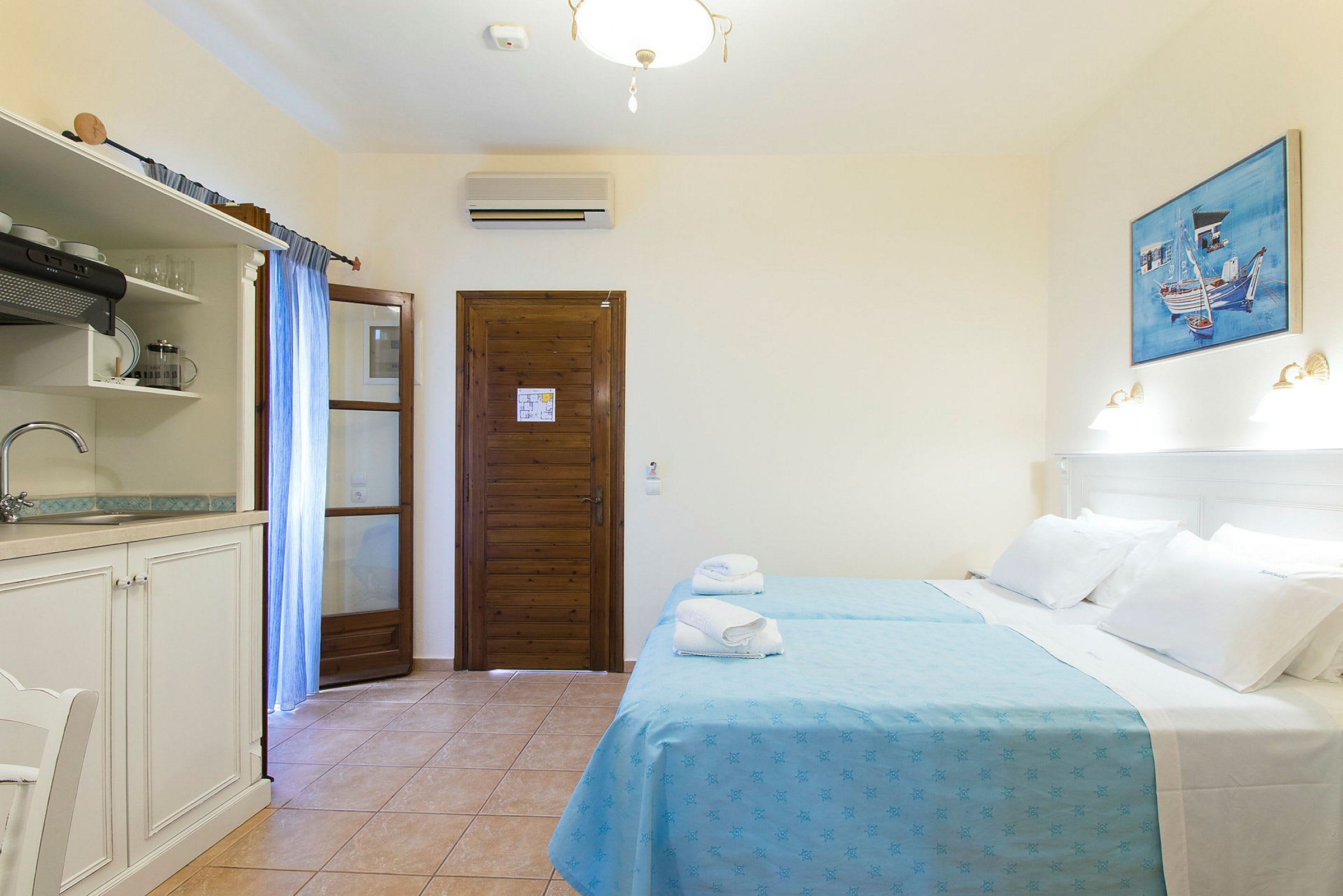 Ett hotellrum med två bäddar klädda i blå sängkläder, en träbeklädd dörr, ett mindre kök med diskho och en målning på väggen. (AI-genererad text)