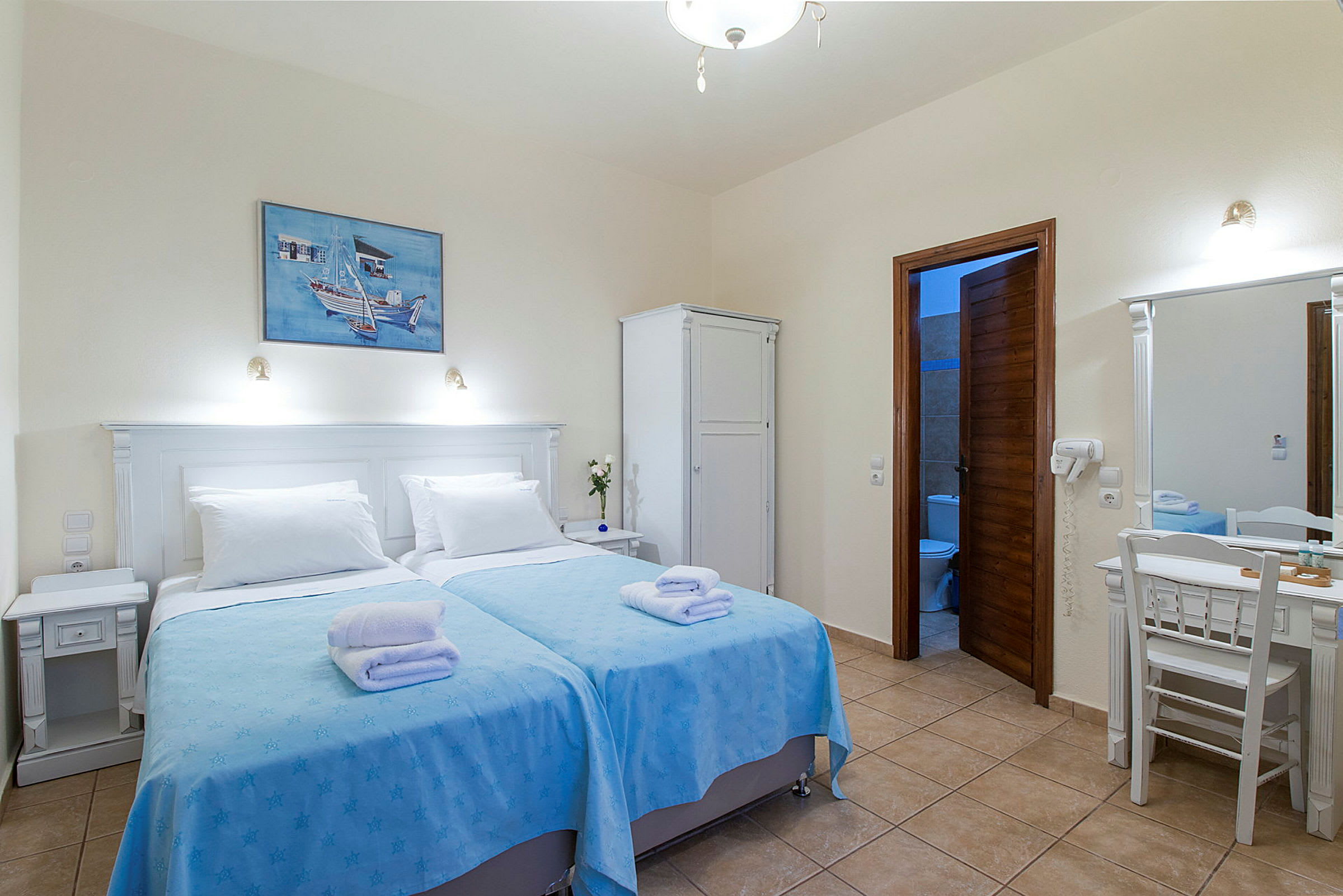 Ett hotellrum med två enkelsängar klädda med blå överkast och vita kuddar, en garderob, ett skrivbord med spegel och ett badrum med öppet dörr. (AI-genererad text)