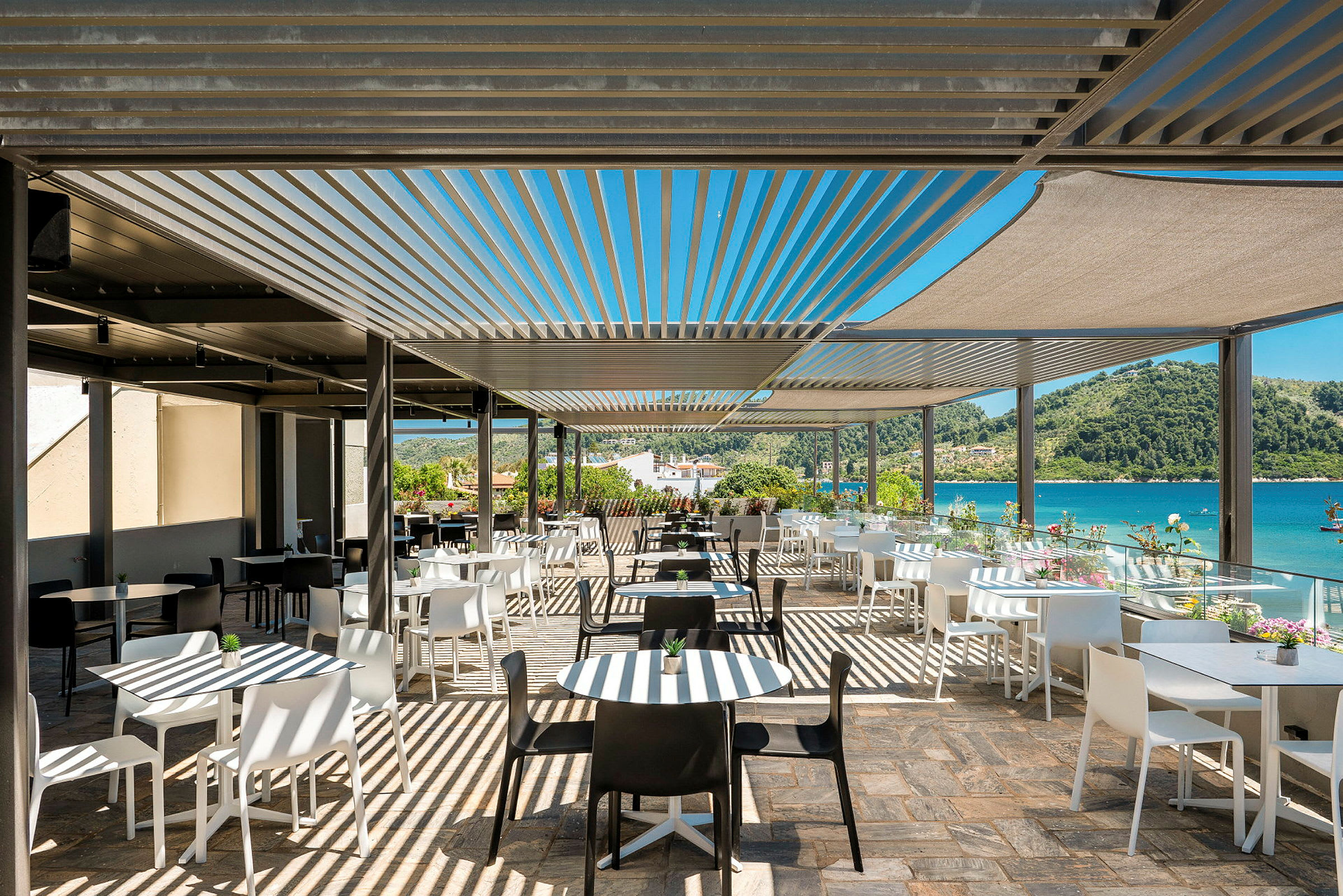 En utomhusrestaurang med vita och svarta stolar och bord under solskydd, med utsikt över havet och omgivande grönska. (AI-genererad text)