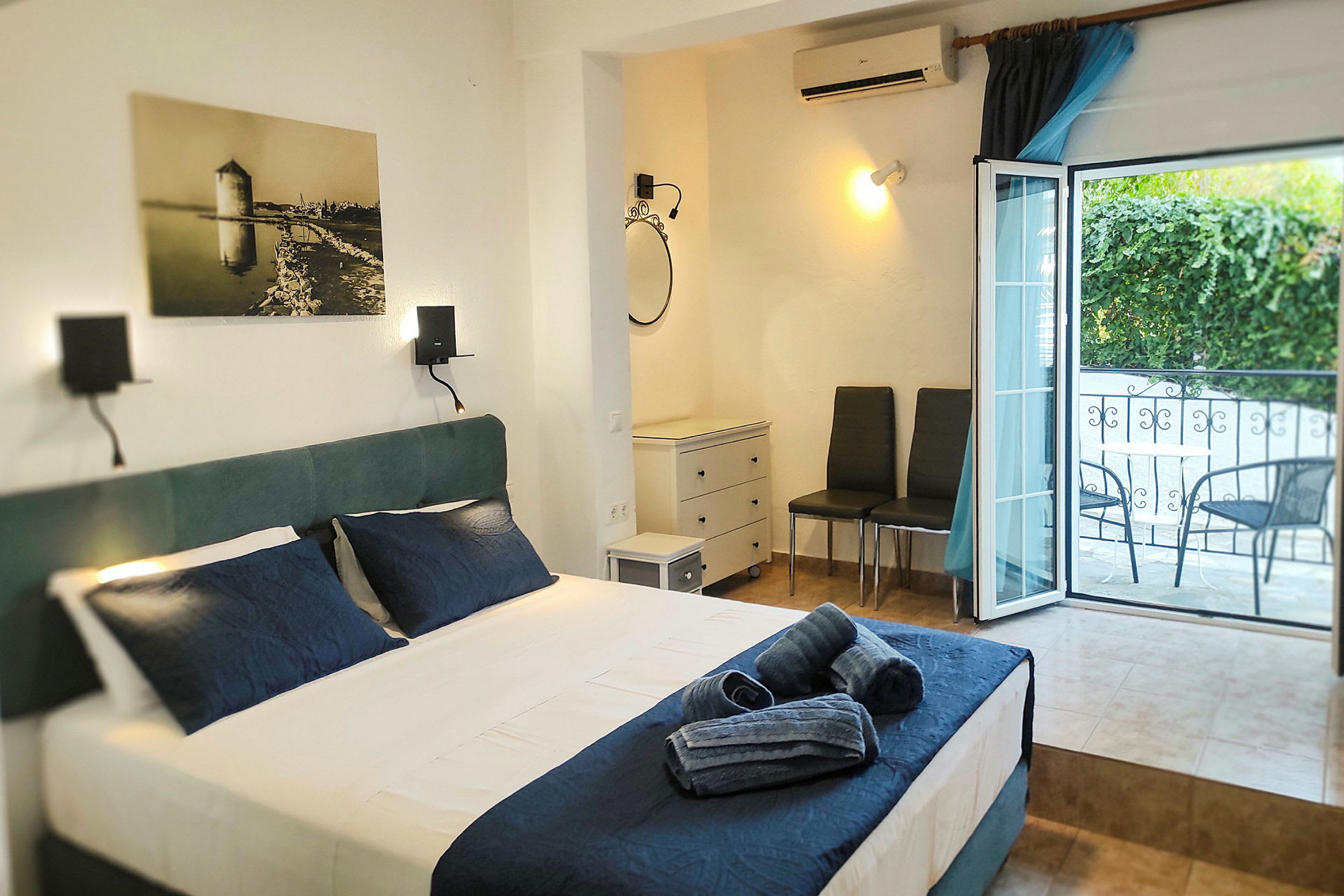 Ett hotellrum med en bäddad säng och mörkblå lakan, ett öppet fönster mot balkongen, och möbler inklusive två stolar och en byrå. (AI-genererad text)