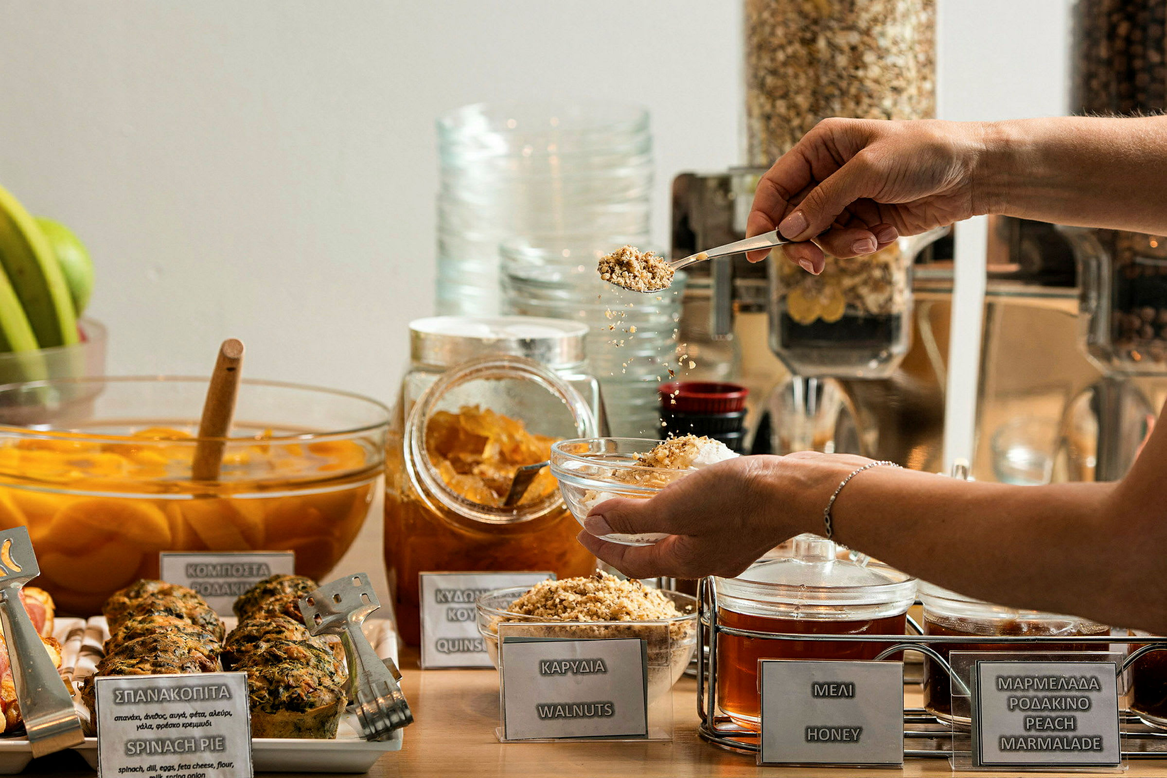 En frukostbuffé med olika val som honung, nötter och marmelad. En person håller en skål och strör nötter över den. (AI-genererad text)