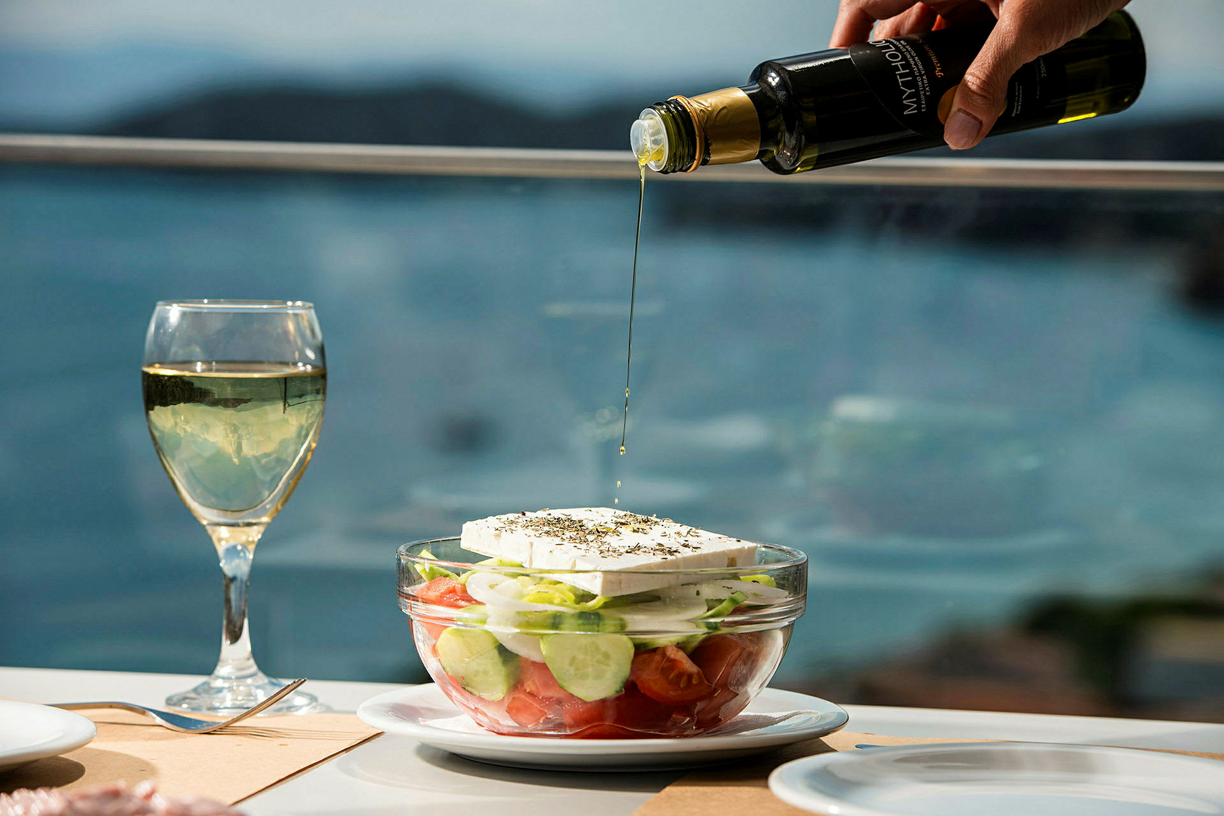 En grekisk sallad i en glasskål vid ett dukat bord. En hand häller olivolja över salladen. (AI-genererad text)